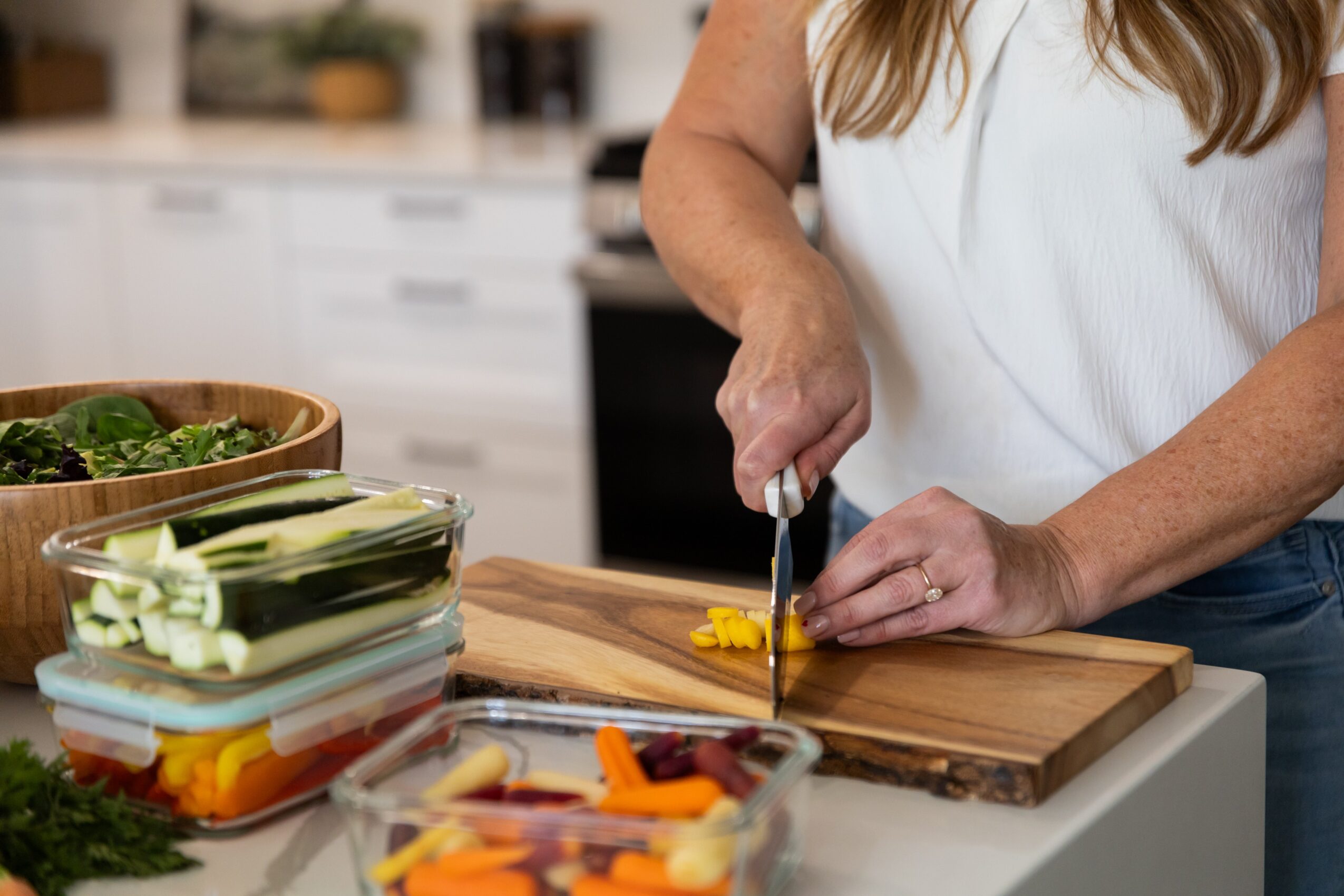 Person chopping vegetables on a wooden cutting board in a kitchen, with containers of sliced vegetables nearby.