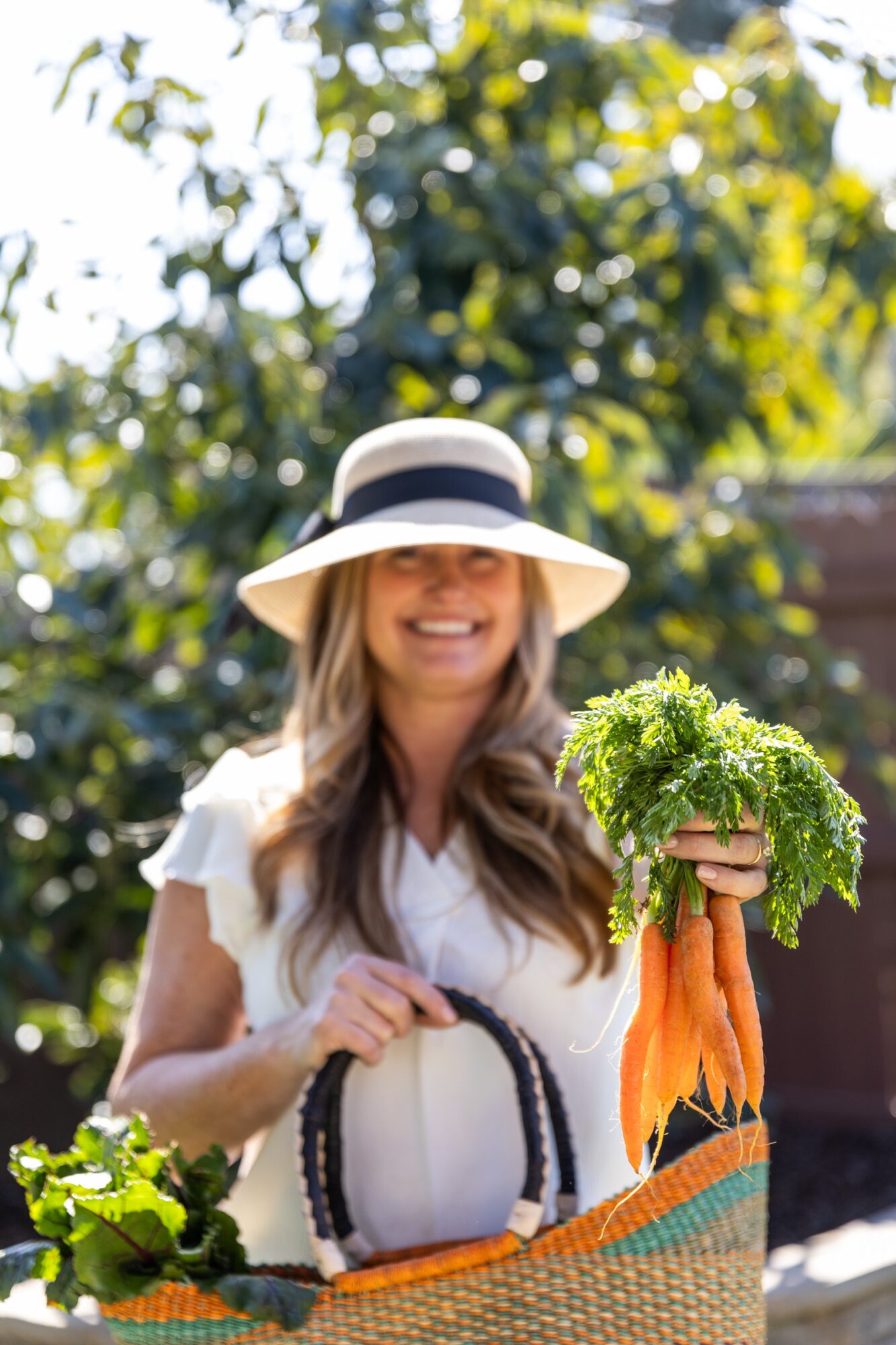 Woman holding a bunch of carrots outdoors, smiling, wearing a wide-brimmed hat and white top, with greenery in background.