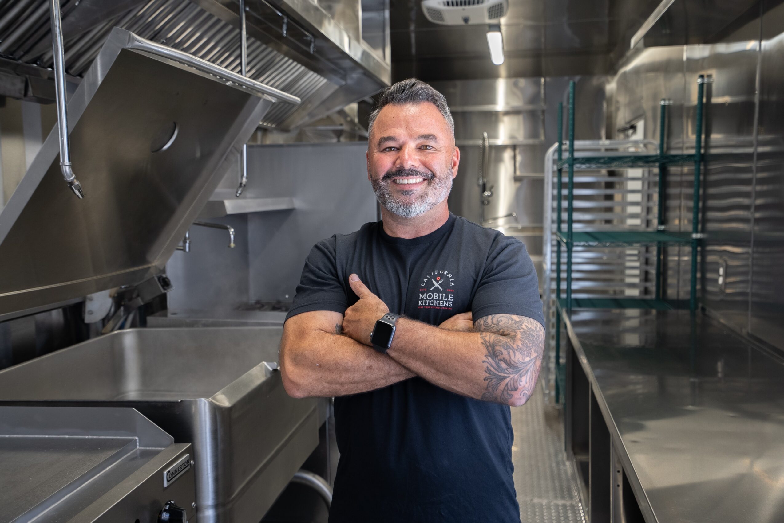 Smiling man with tattoos and a beard in a commercial kitchen, arms crossed, wearing a black T-shirt.