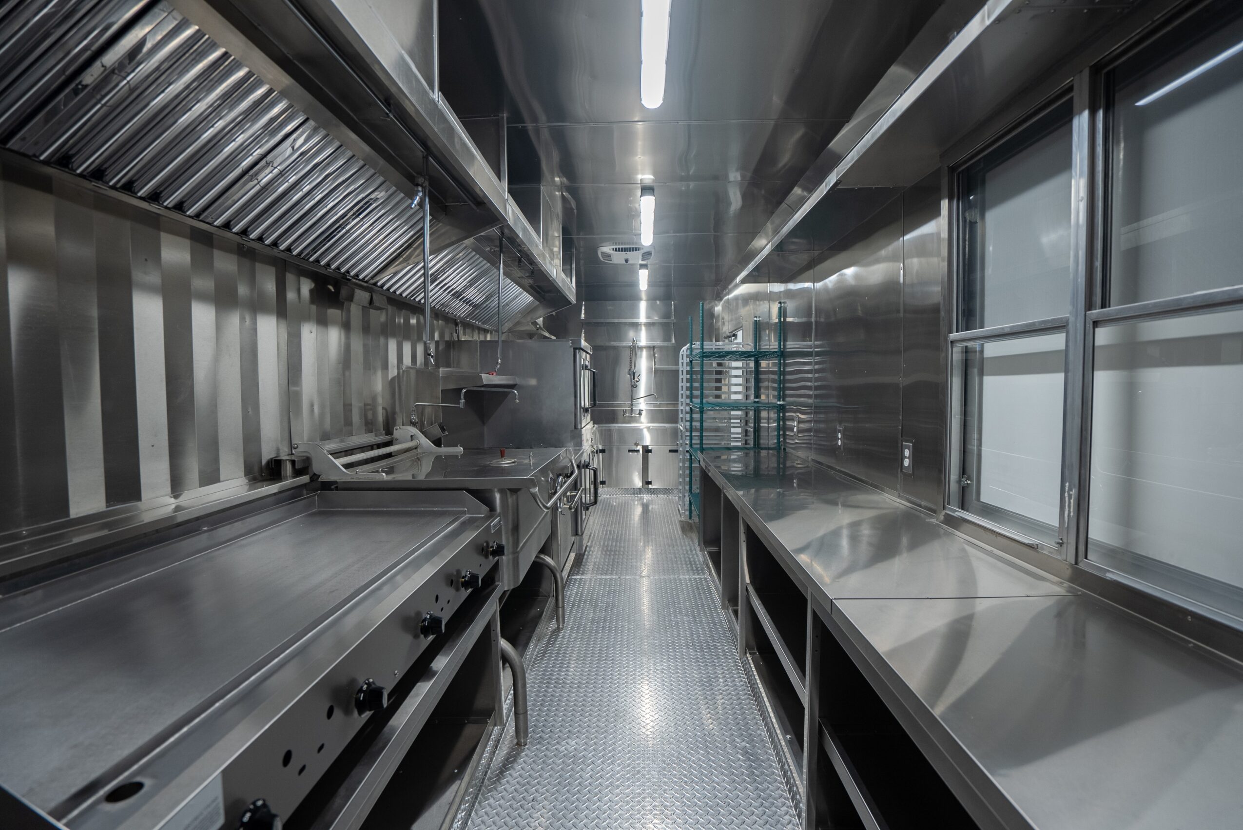 Empty stainless steel kitchen corridor with reflective surfaces and large windows on the right side.