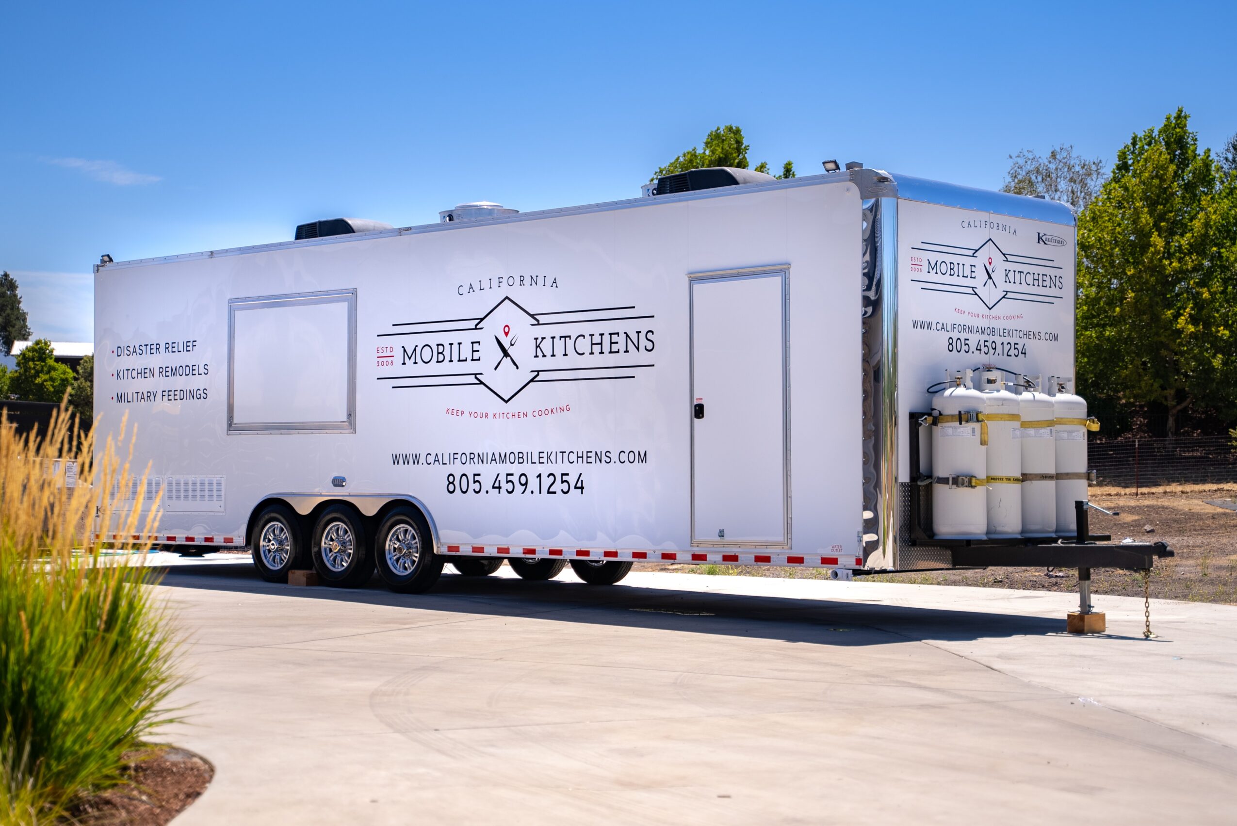 Mobile kitchen trailer parked outdoors with trees and blue sky in background, displaying company logo and contact information.
