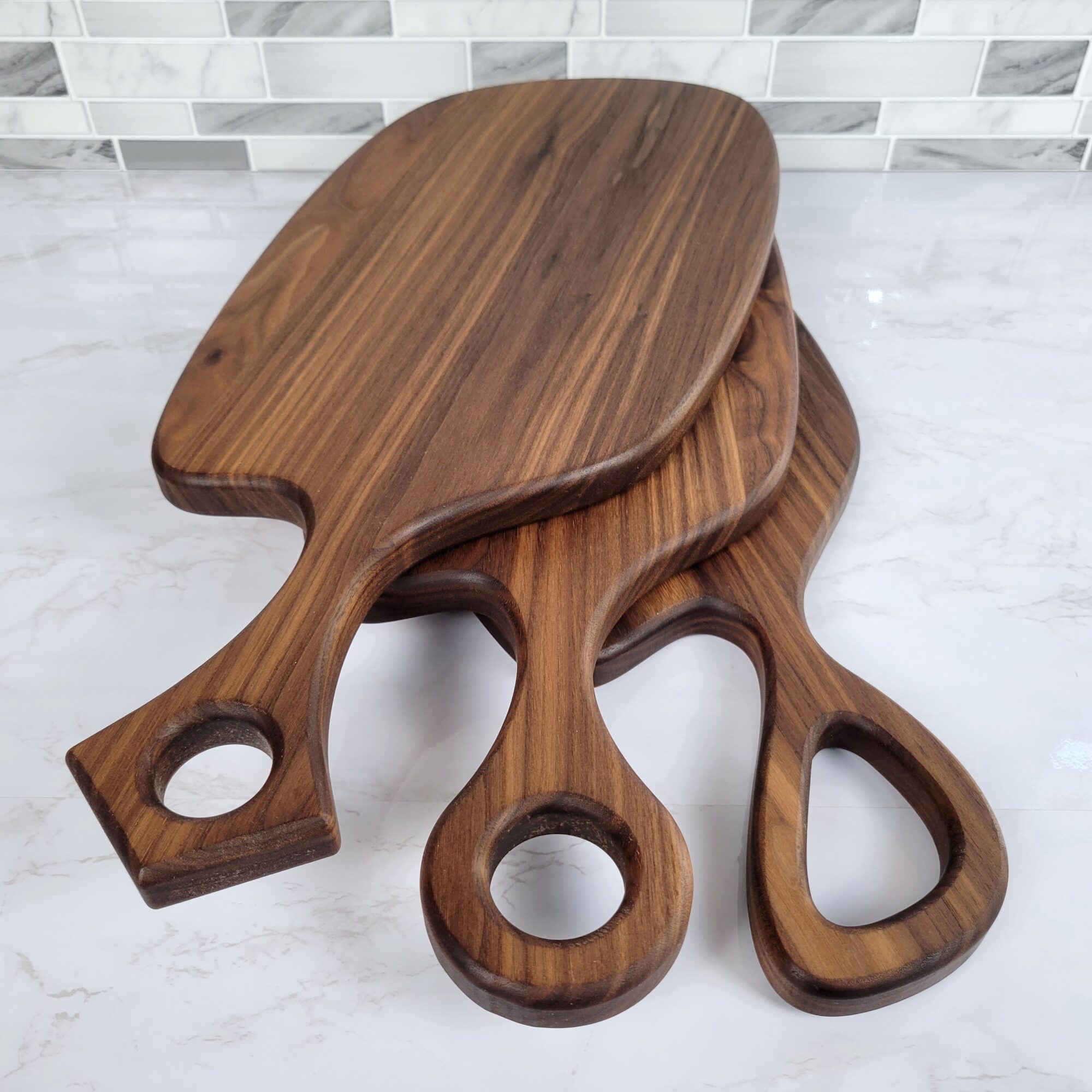 Three wooden cutting boards stacked, with handles and circular holes, on a marble surface with a tiled wall background.