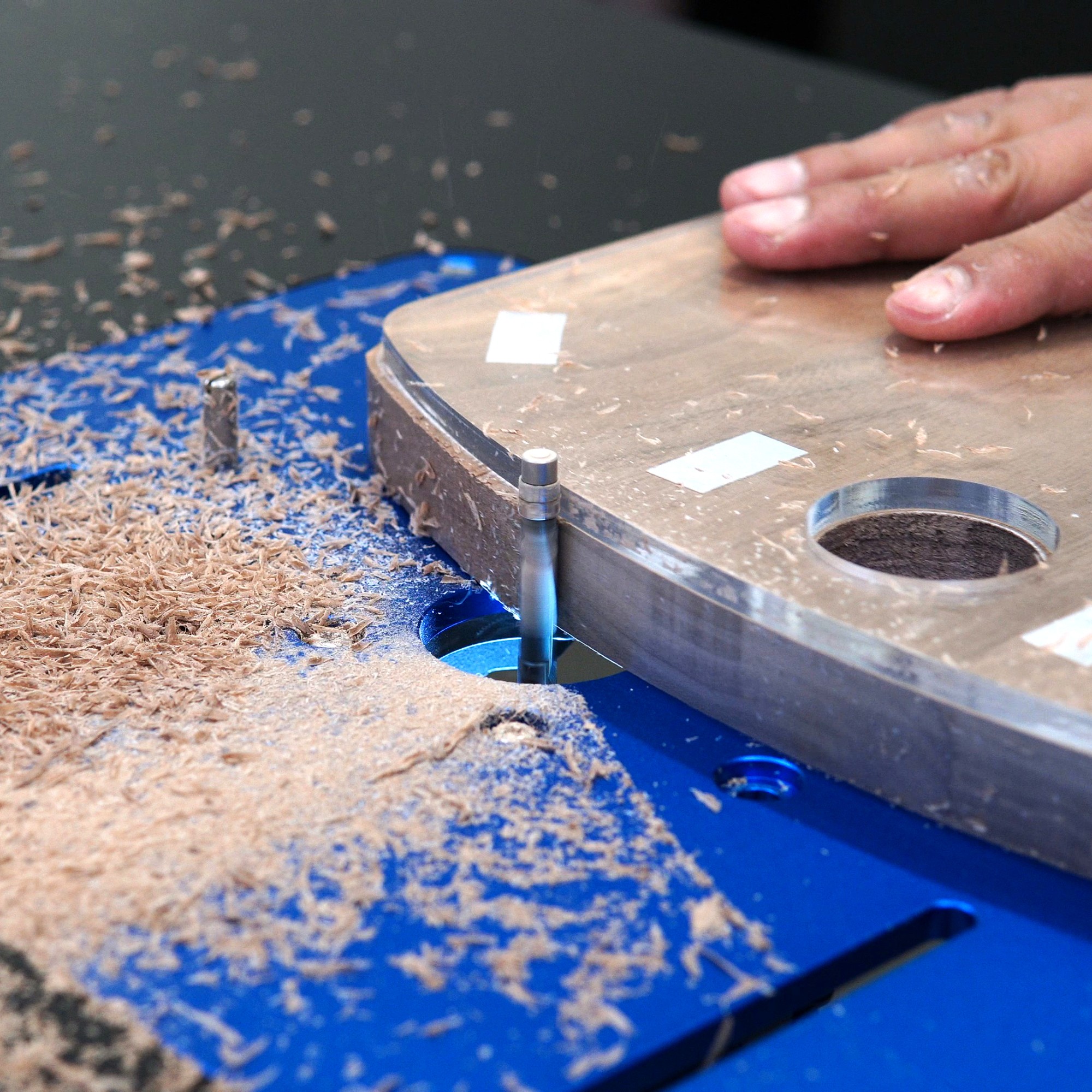 Person's hand guiding a router on a wooden piece, with wood shavings on a blue surface.