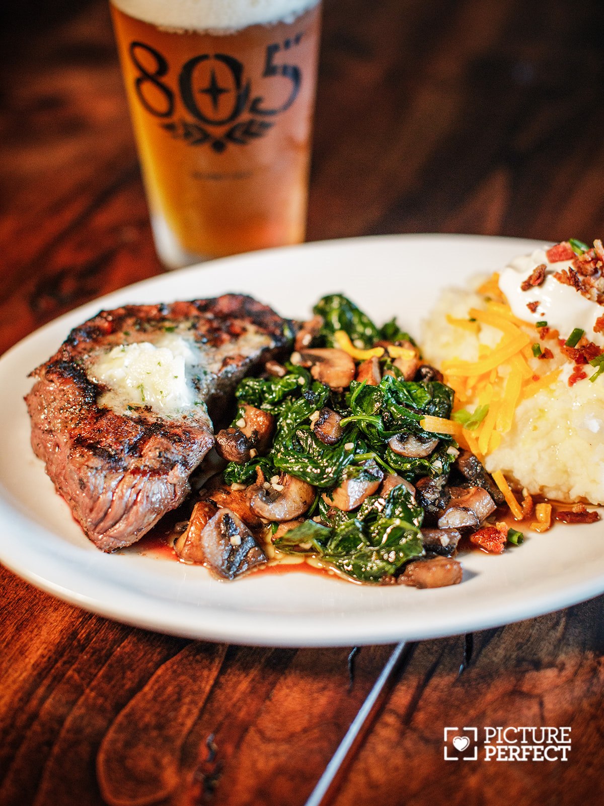 Steak with sautéed spinach, mashed potatoes, and a glass of beer in the background on a wooden table.