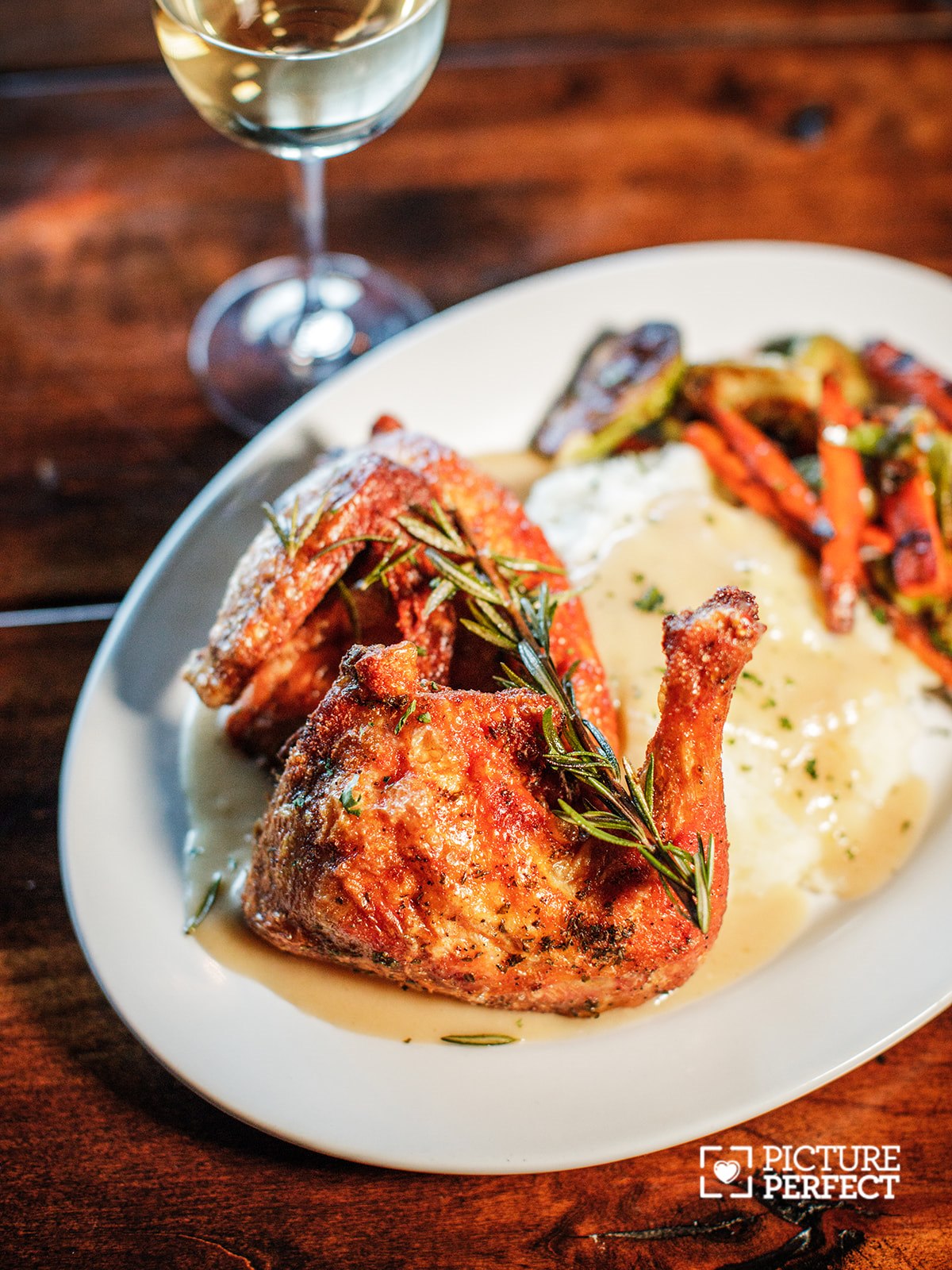 Fried chicken with herbs, mashed potatoes, and vegetables on a white plate, with a glass of white wine in the background.