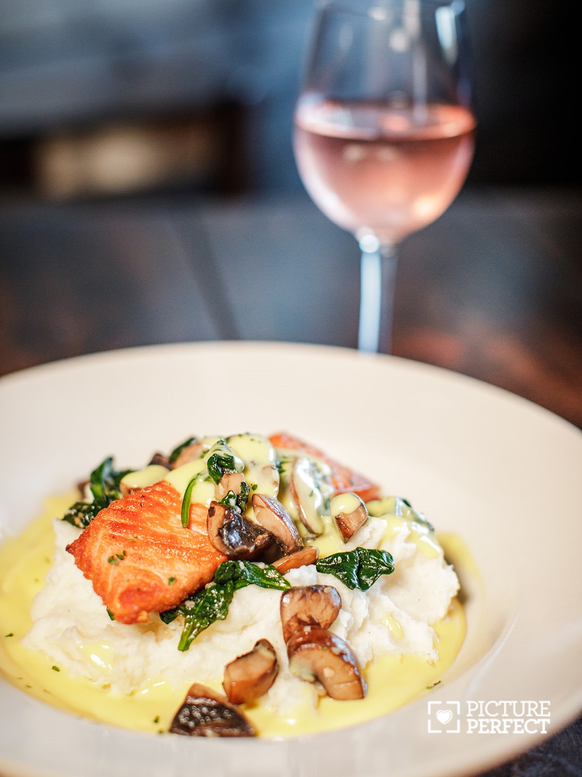 Plate of creamy mashed potatoes topped with salmon, mushrooms, and greens, with a glass of rosé wine in the background.