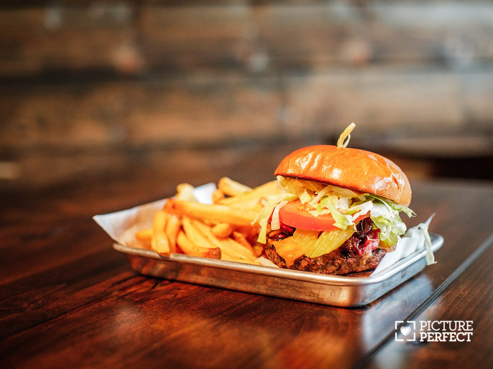 Burger with lettuce, cheese, and patty, served with fries on a metal tray, on a wooden table, blurred background