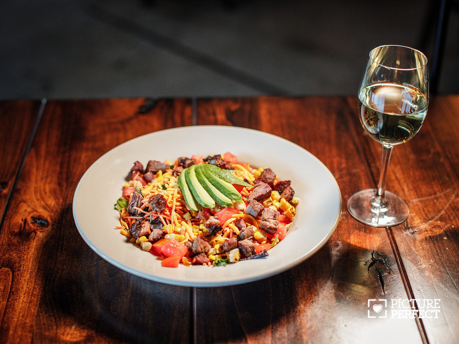Plate of colorful food with avocado slices on top, next to a glass of white wine on a wooden table.
