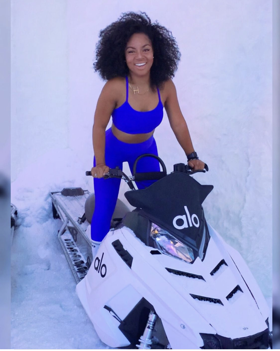 Woman in blue athletic outfit smiling, standing next to a white snowmobile in snowy environment.