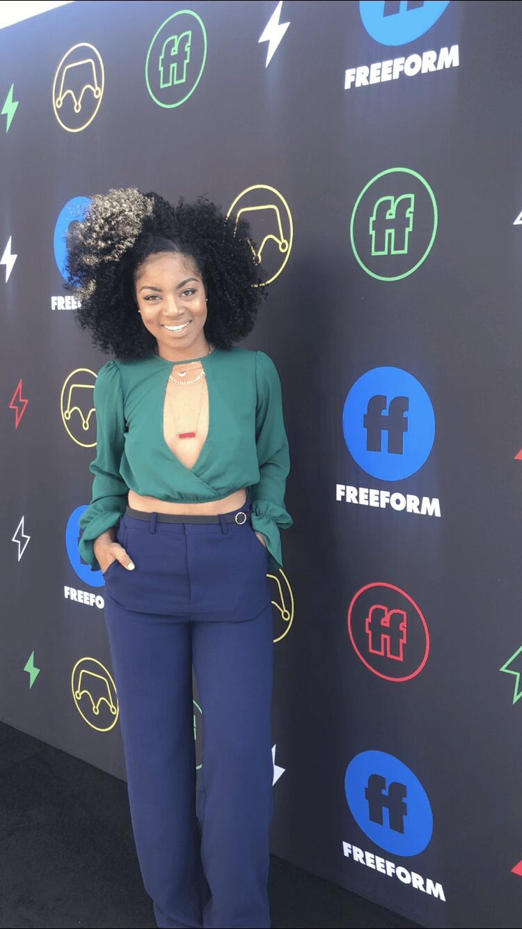 Young woman with curly hair in a bun, smiling, wearing a green top and blue pants, standing against a black background with logos.