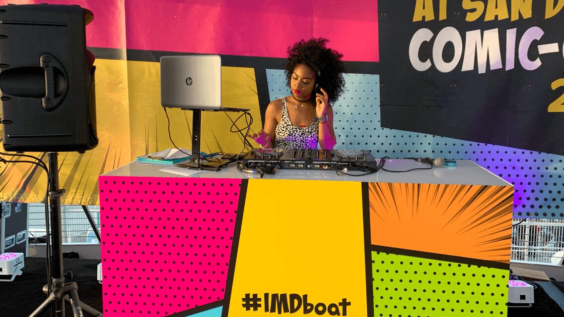 A DJ with curly hair and colorful clothing performs at a colorful booth with a computer and DJ equipment, at a comic convention.