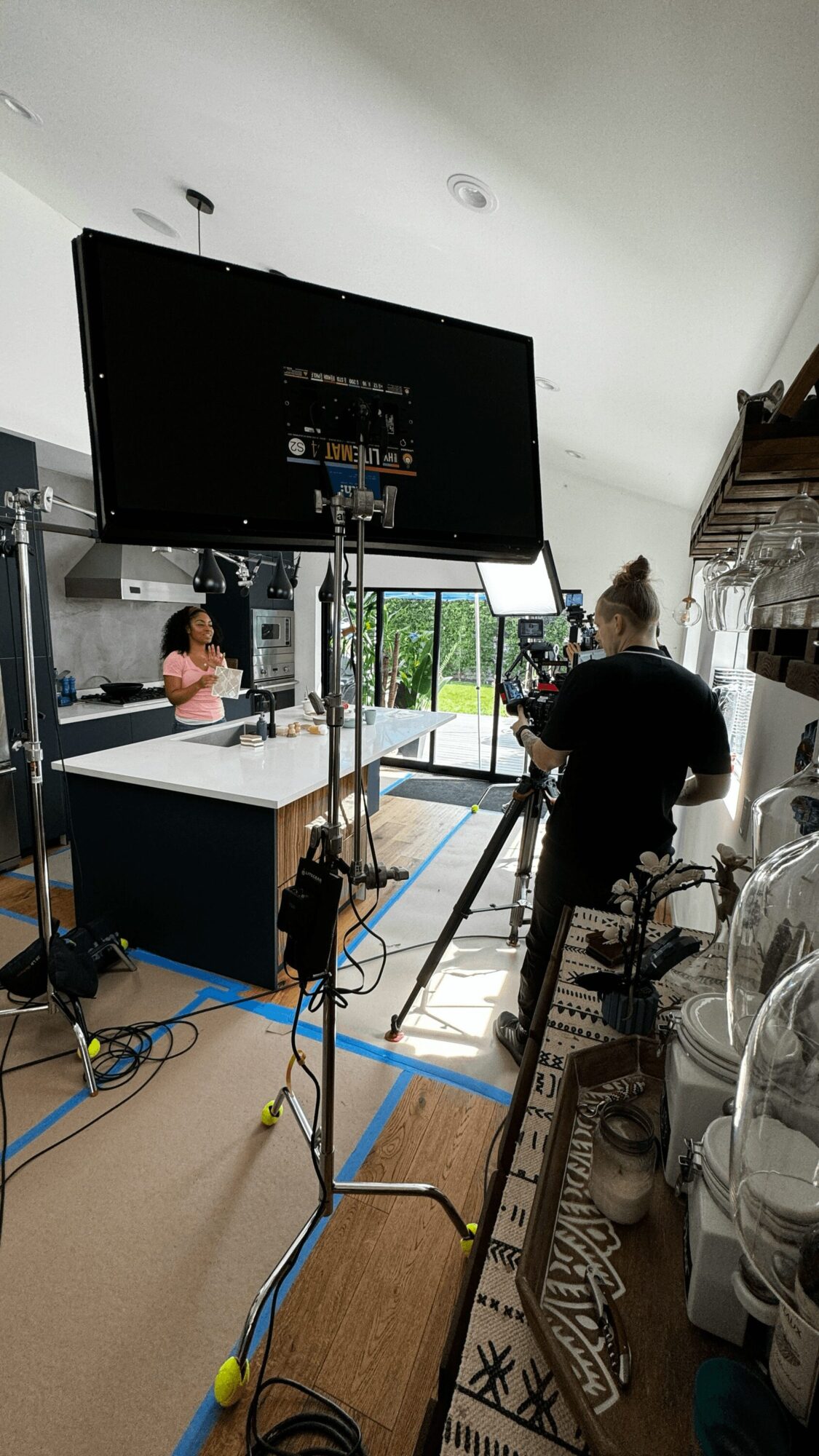 Person filming a woman speaking at a table in a bright room with large windows, equipment setup visible.