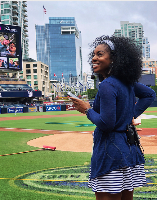 Smiling woman with curly hair in a blue jacket and striped dress at a baseball stadium, holding a phone, city skyline in background.