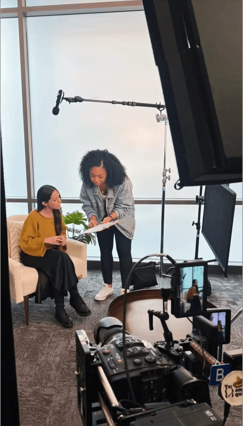 Two women in a room with filming equipment, one sitting on a sofa and the other standing, reading a script.