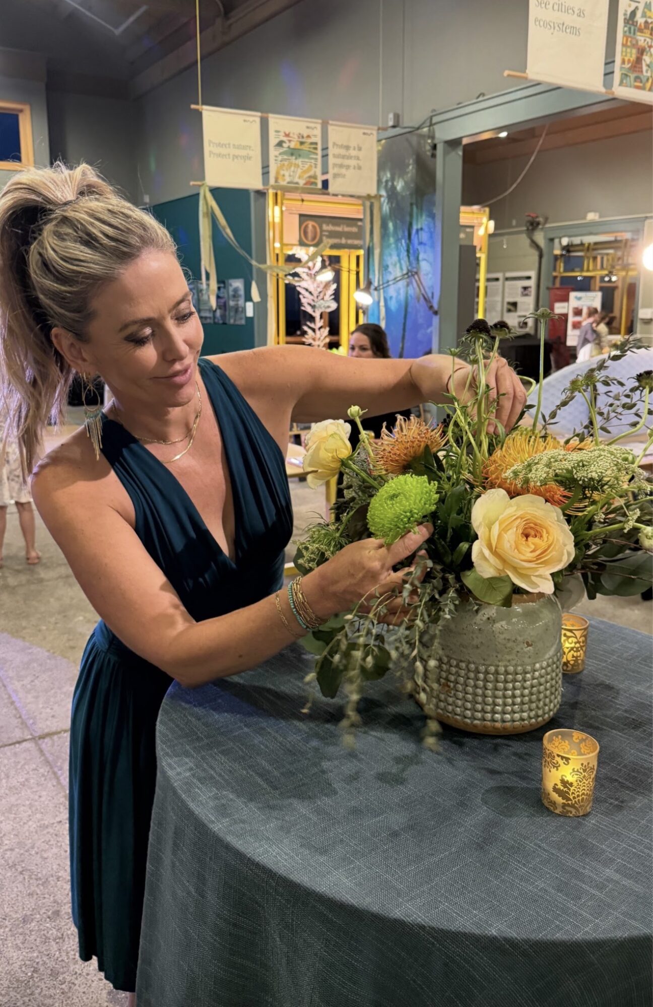Woman arranging a floral display with roses and greenery at a table in an indoor setting.