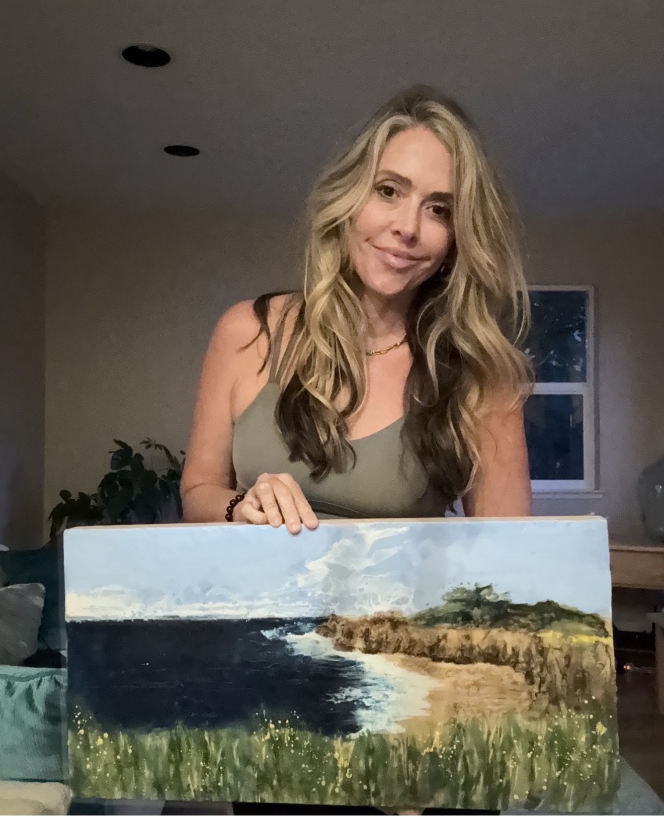 Woman with long blonde hair holding a landscape painting, standing indoors near a window, smiling.