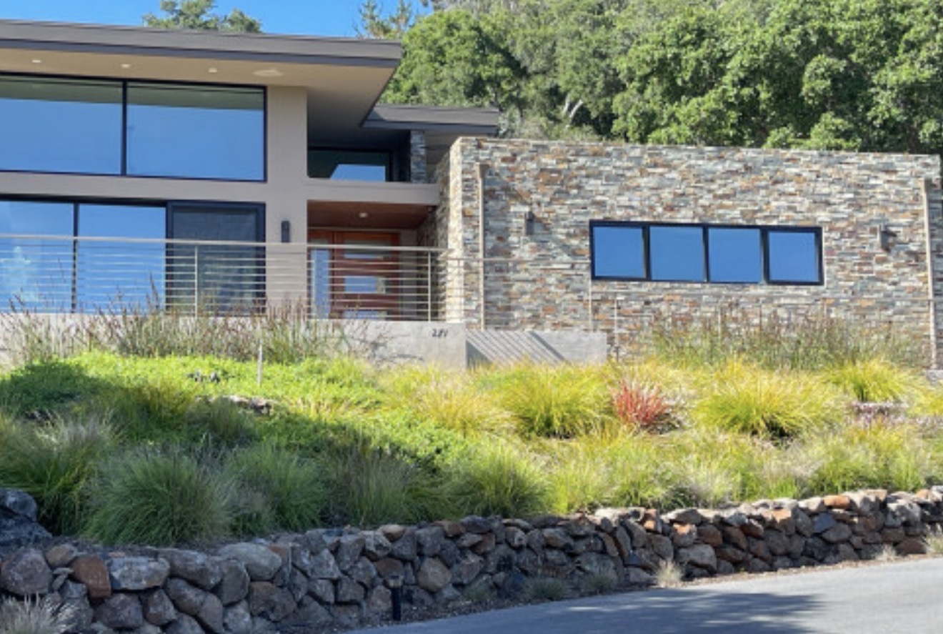 Modern house with large windows, stone wall, and landscaped front yard with grass and plants.