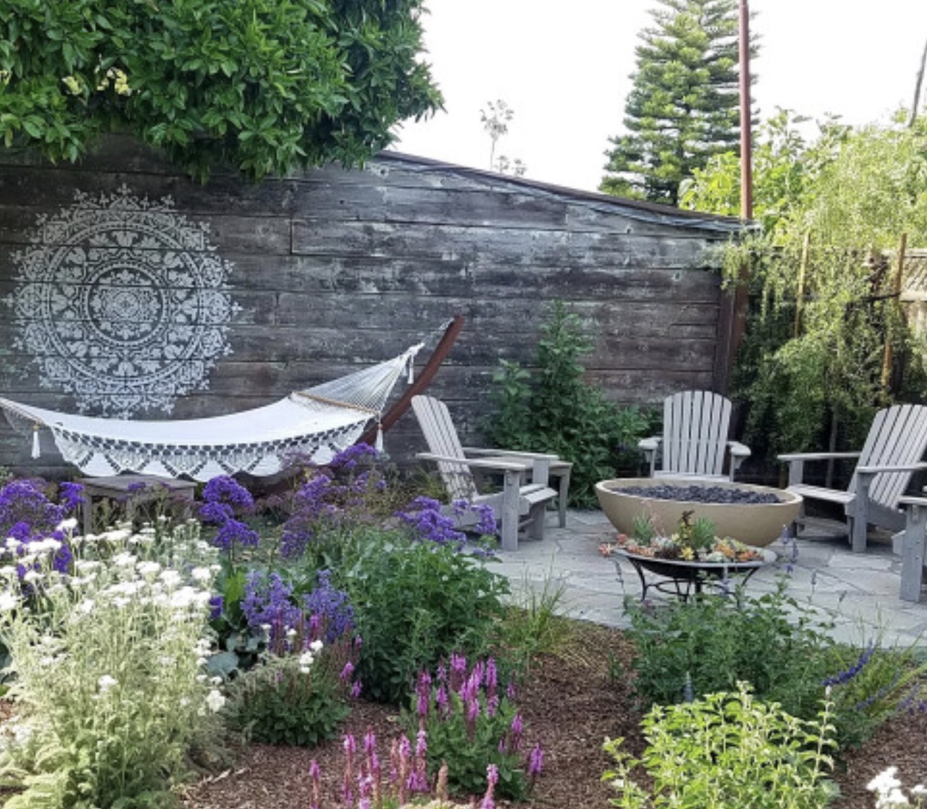 Backyard garden with a hammock, colorful flowers, and outdoor seating area, enclosed by a wooden fence and greenery.