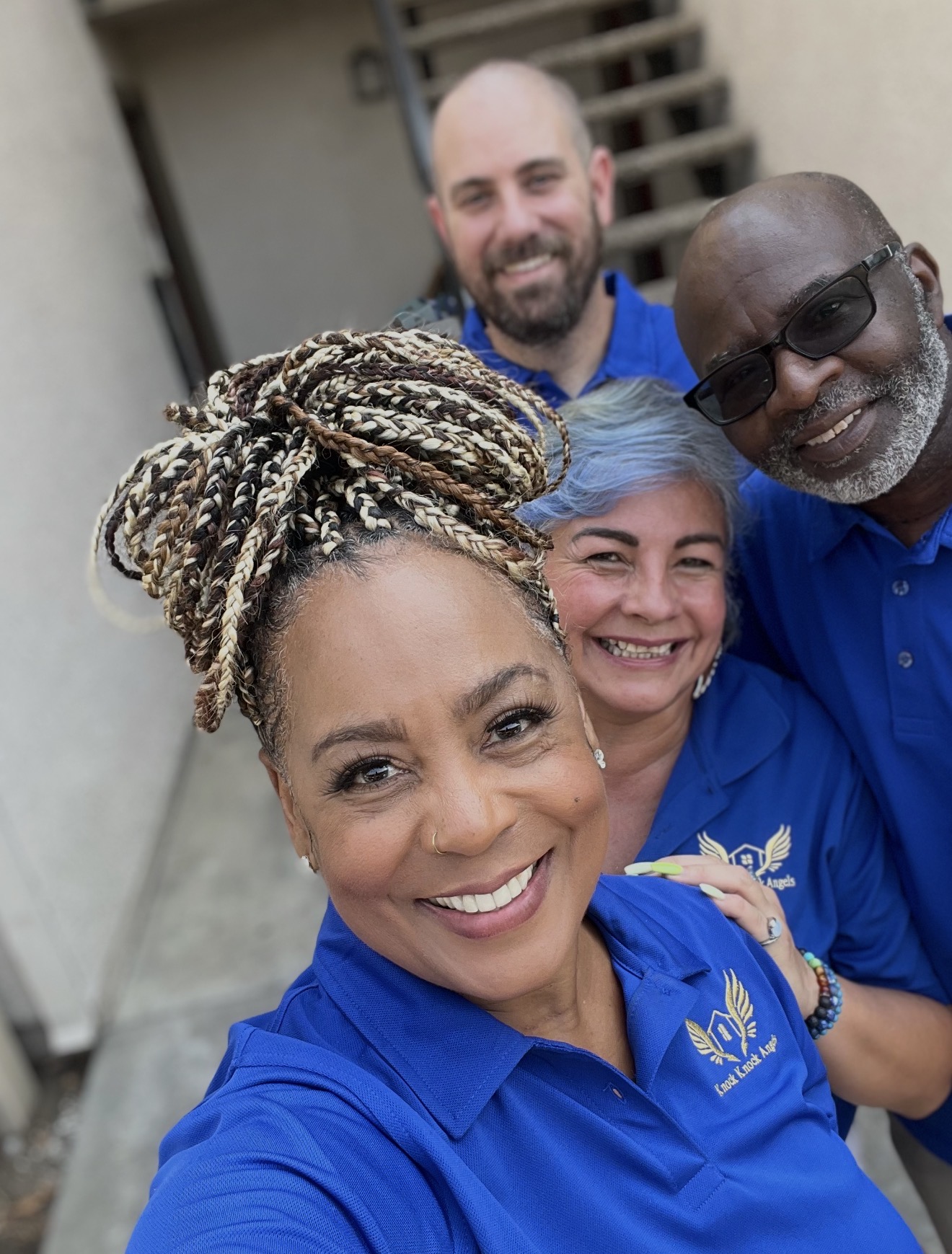 Four smiling people in blue shirts taking a selfie outdoors, with stairs and a building in the background.
