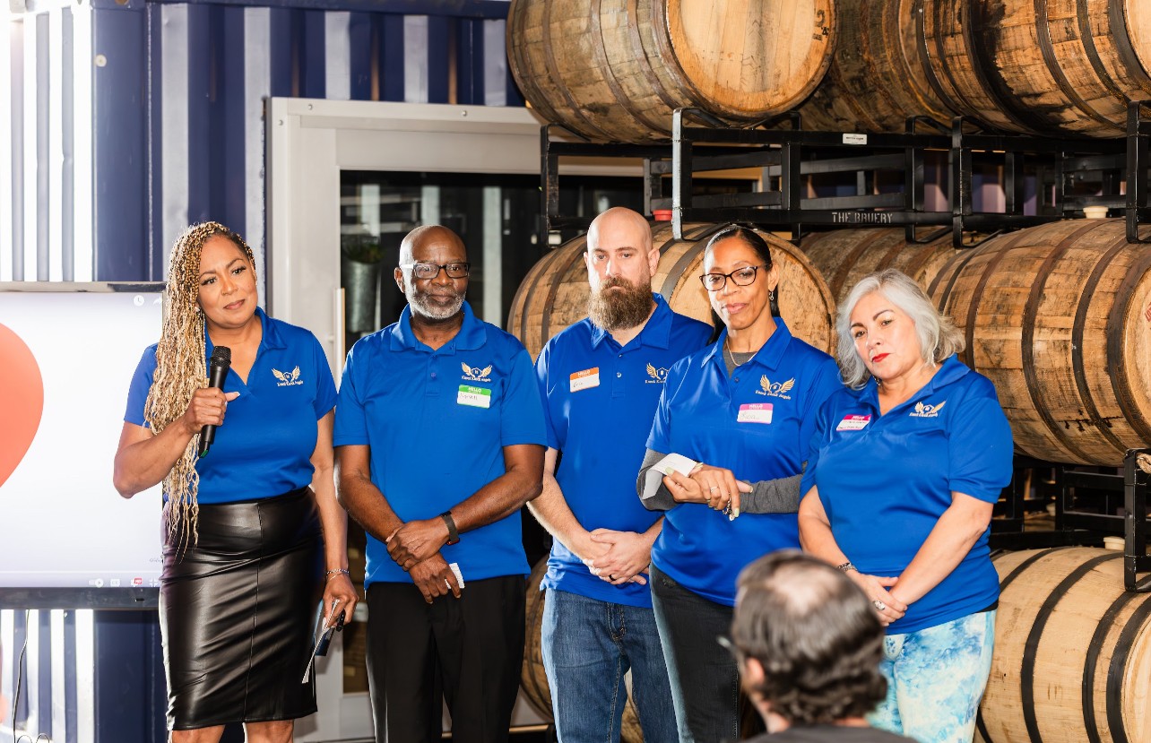 Group of five people in blue shirts standing in front of barrels, listening to a person speaking, in an industrial setting.