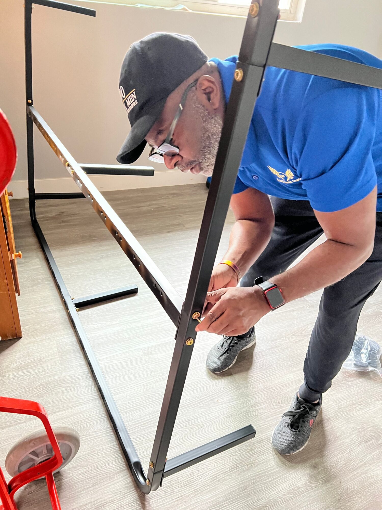 Person in blue shirt and black pants writing on a whiteboard, with a red object nearby, in a room with wooden floor.