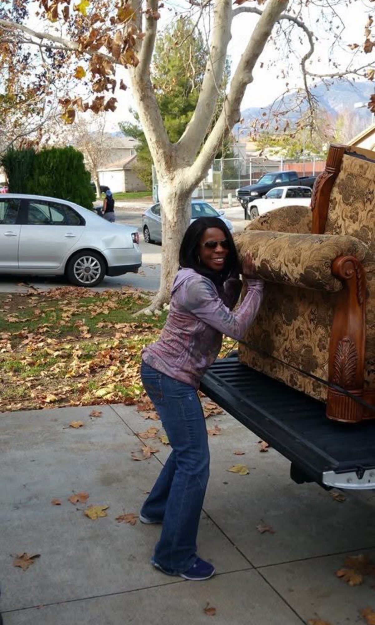 Person standing outside near a tree, holding a large object on a truck bed, with cars and houses in the background.