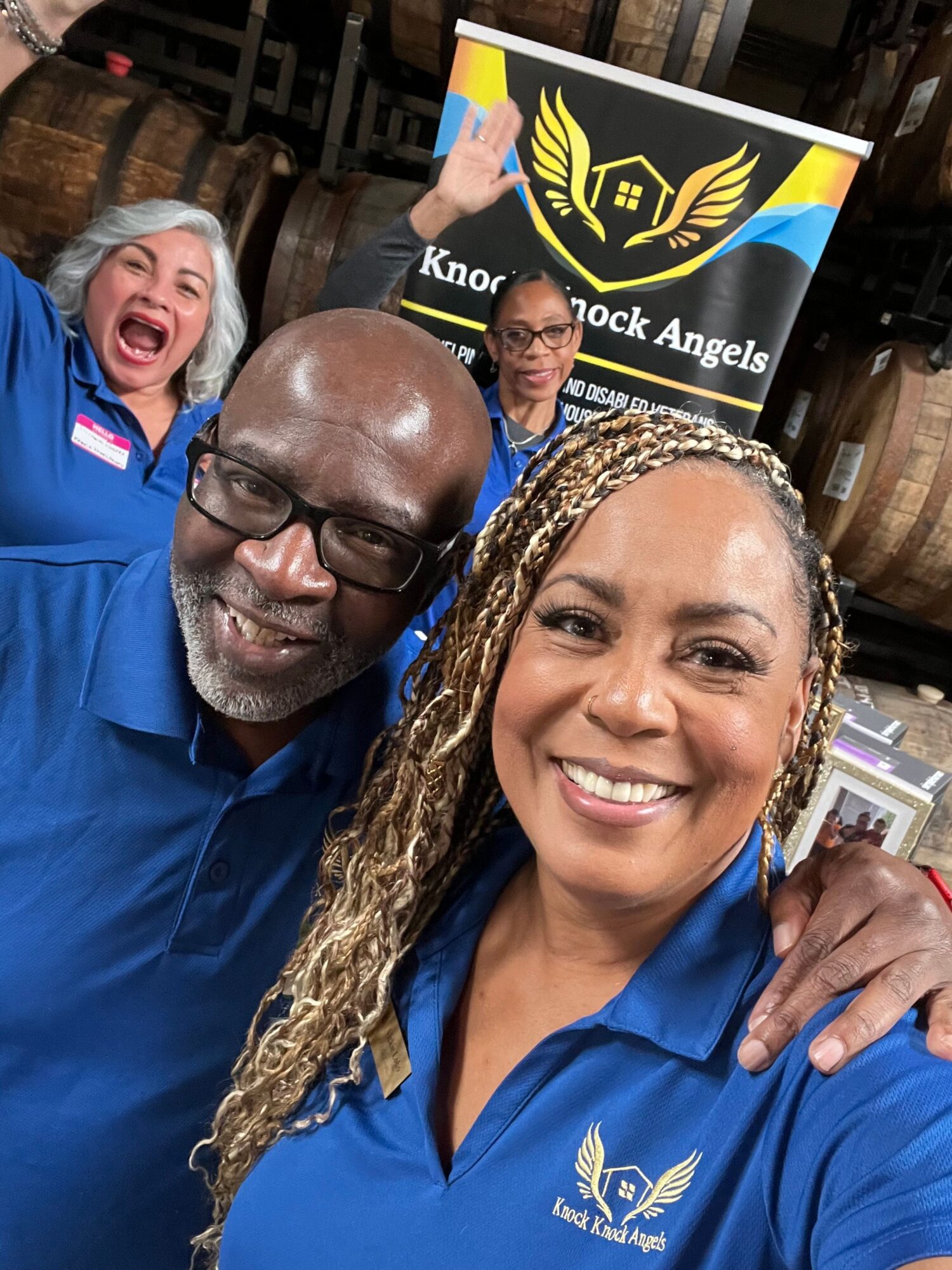 Two smiling women and a man in blue shirts, with a Knorrock Angels sign in the background.