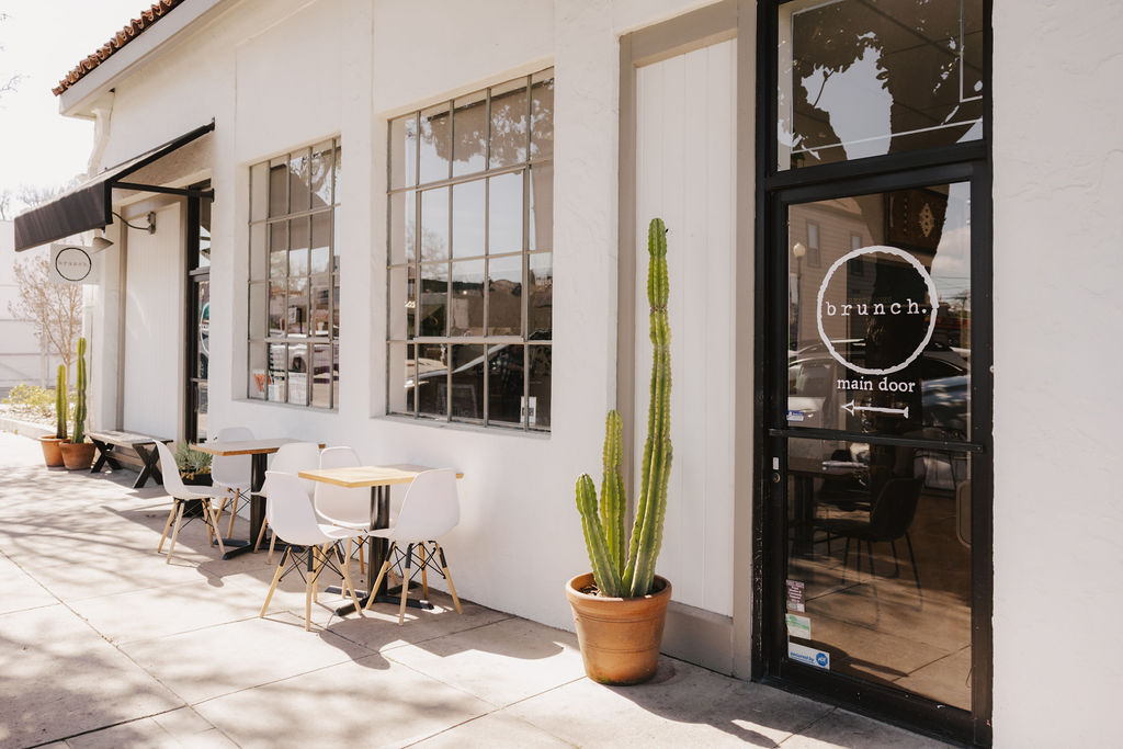 Exterior of a building with large windows, outdoor seating, and a tall potted cactus near the entrance.