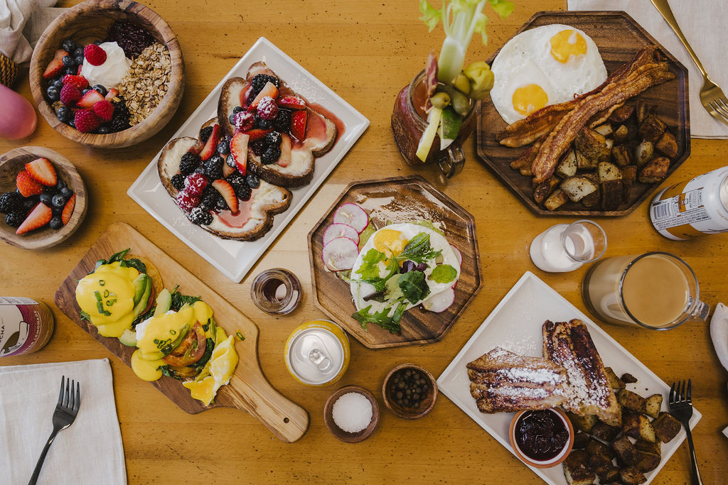 Assorted desserts and drinks on a wooden table, including cakes, pastries, fruit, and beverages, with utensils and small bowls.