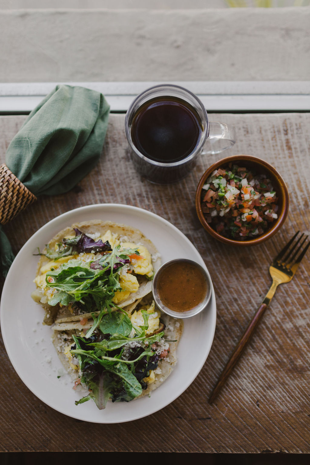 Plate with sandwich and salad, bowl of soup, glass of dark beverage, small bowl of toppings, fork, on wooden table.
