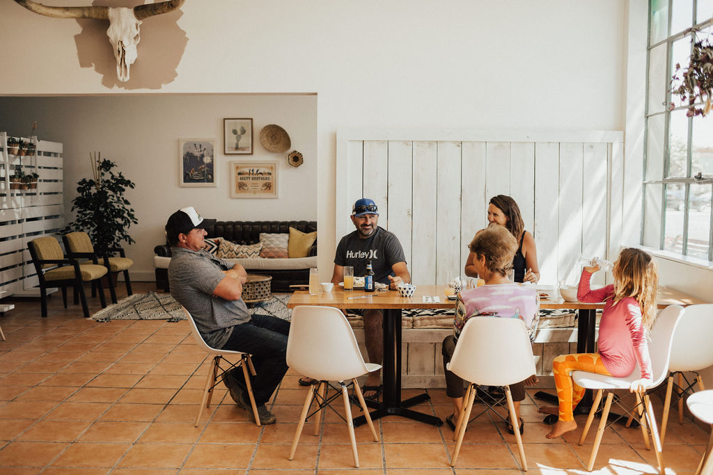 Group of six people sitting around a table in a bright room with large windows, some talking and others listening.