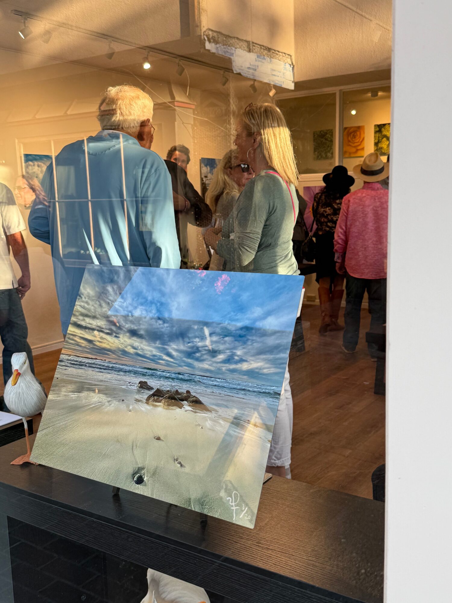 People gather in a room with a painting of a beach scene on a table, reflecting the room's interior and visitors.