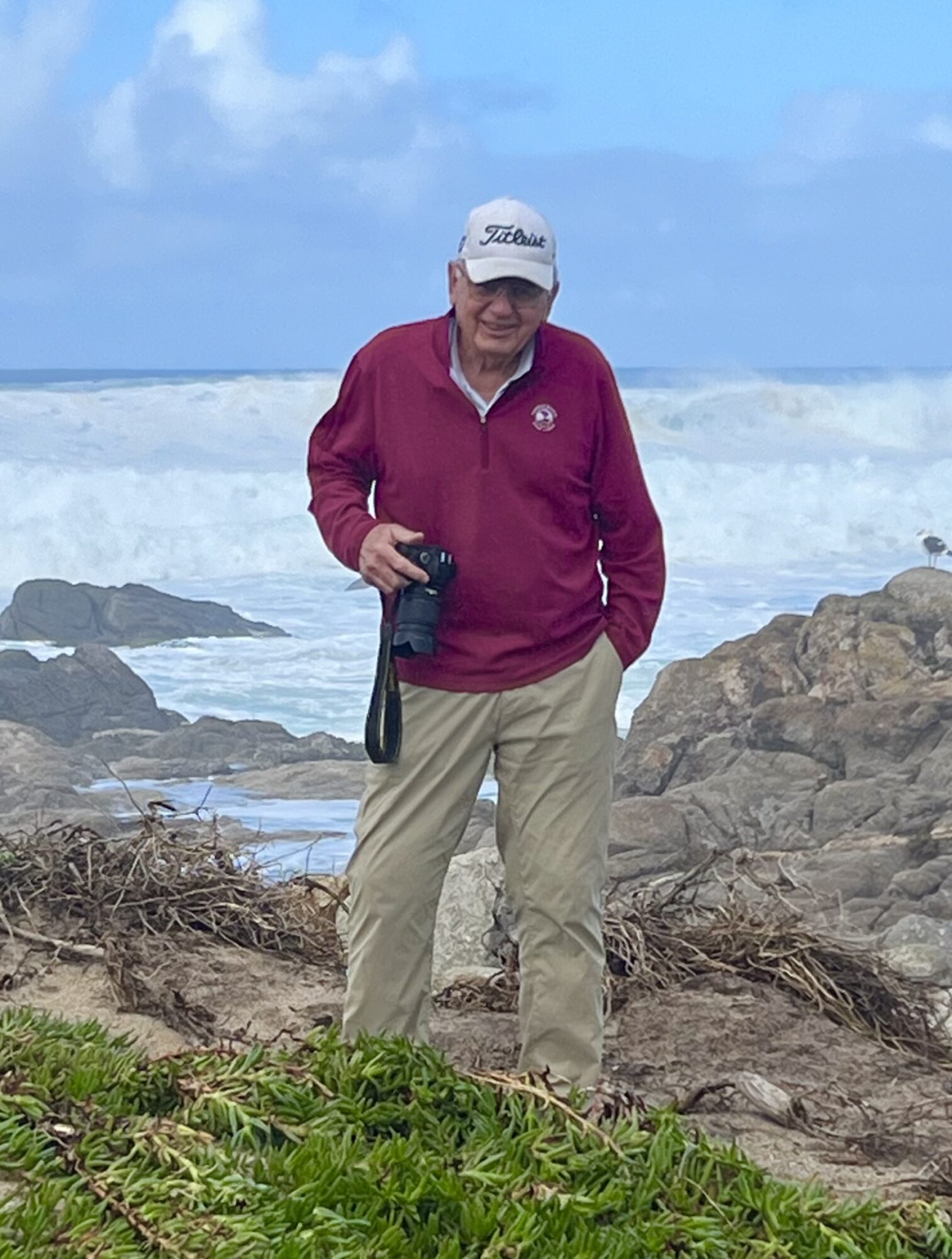 Person standing on rocky beach with ocean waves and blue sky in background, holding a camera, wearing a white cap, red jacket, and beige pants.
