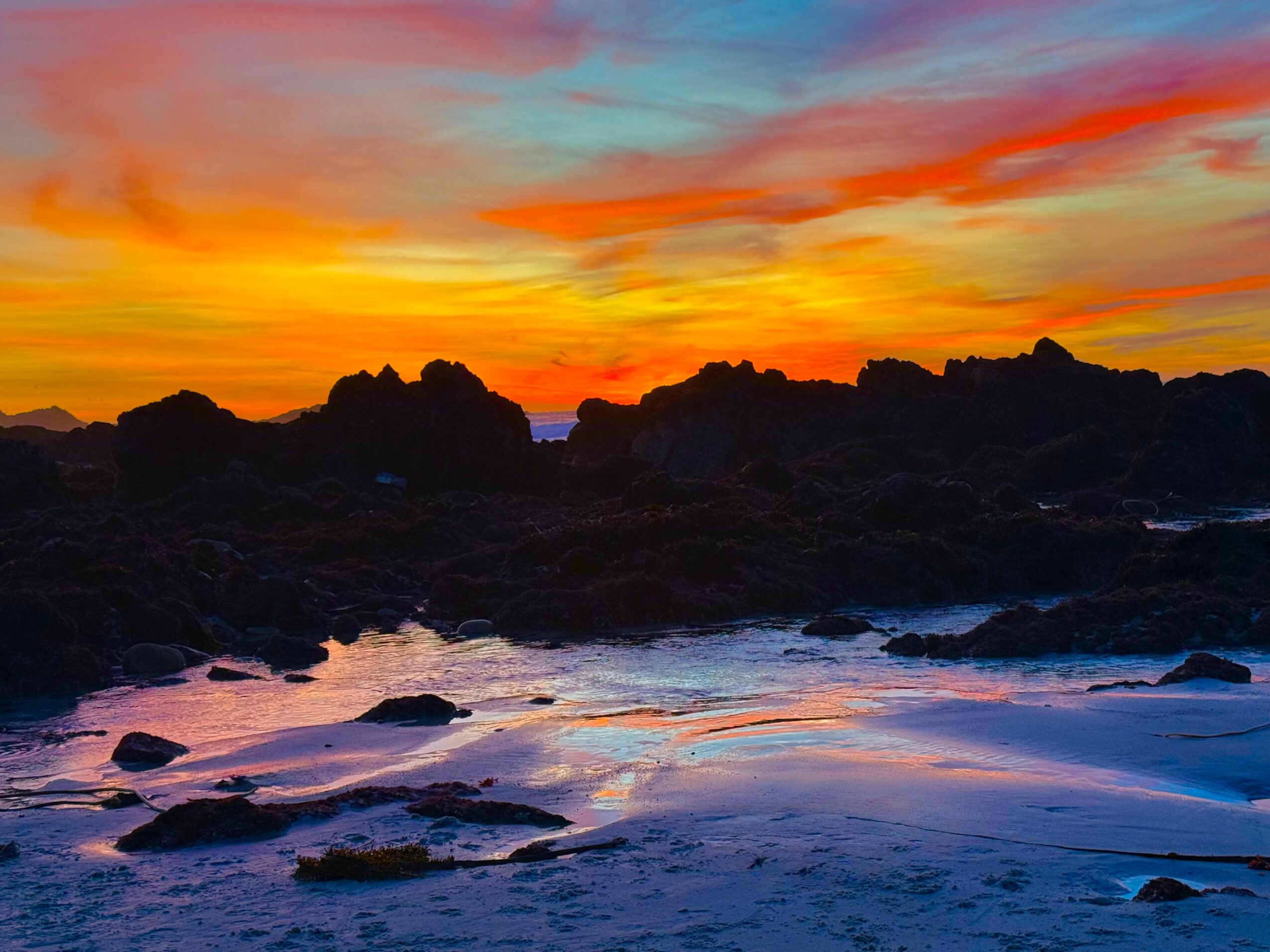 Colorful sunset sky over a rocky shoreline with waves, vibrant orange, pink, and blue hues, and dark rocks in foreground.
