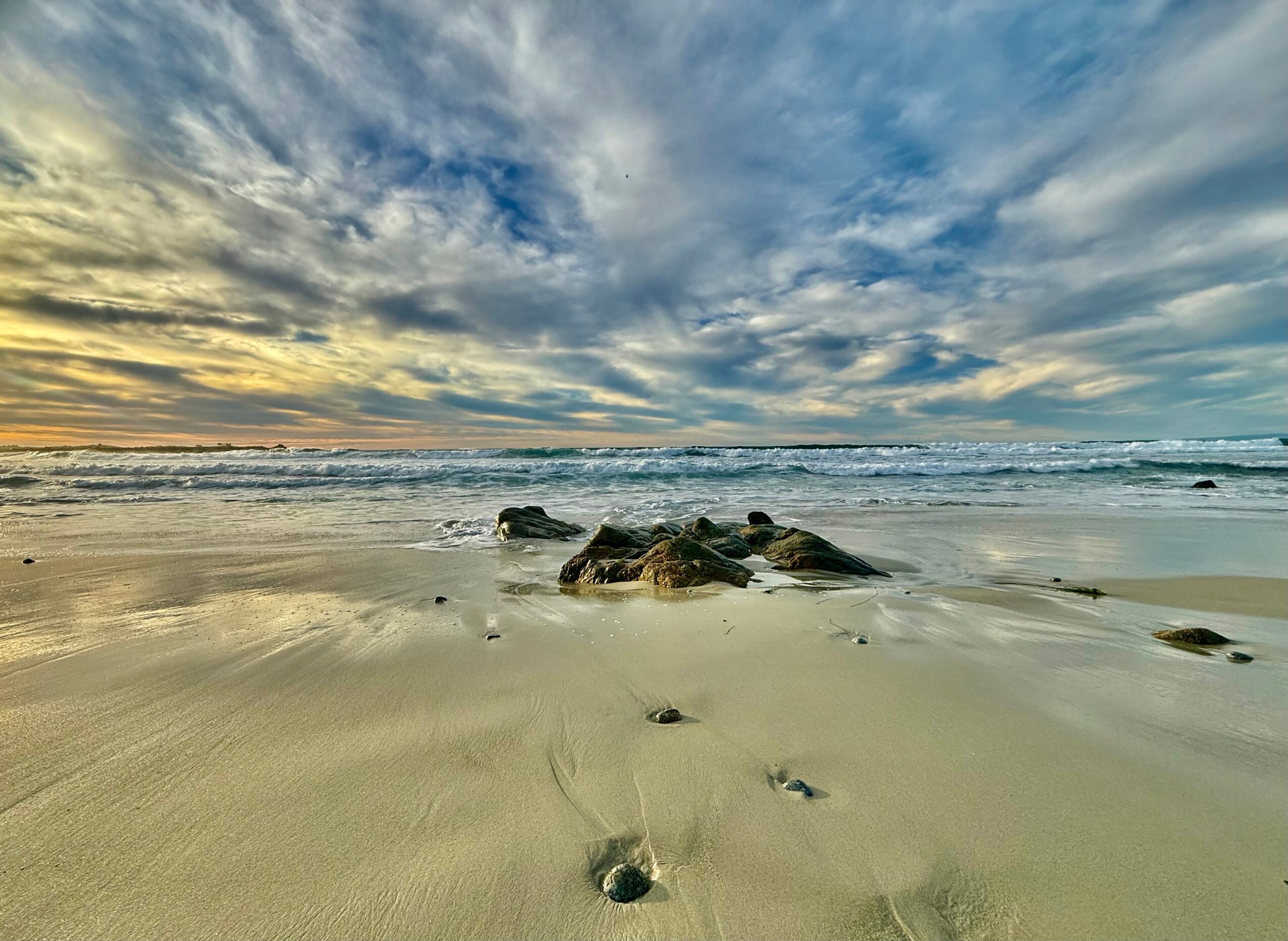 Beach with sand, rocks, and footprints, under a cloudy sky with sunlight near the horizon.