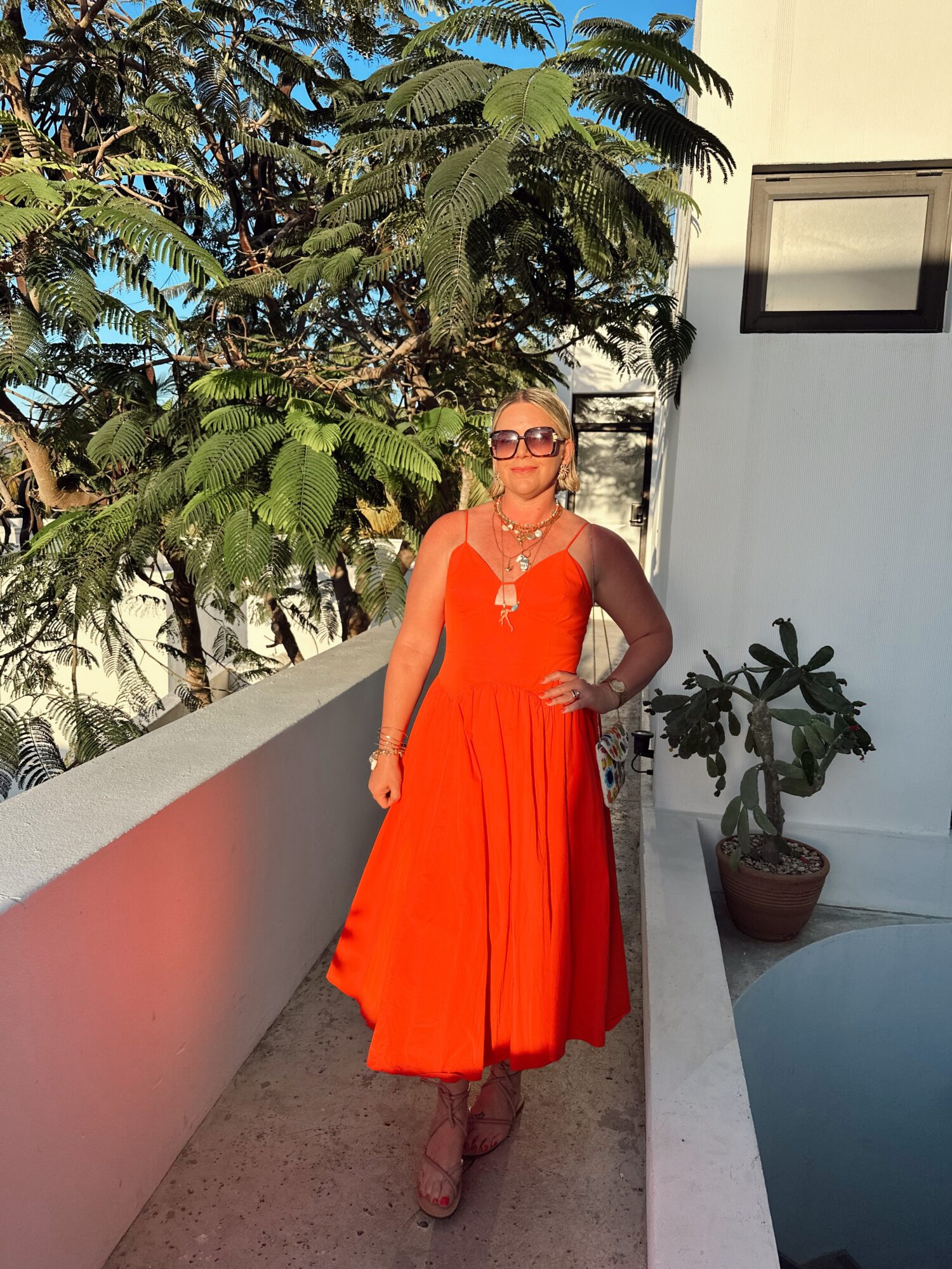 Woman in a bright orange dress and sunglasses standing outdoors near a white wall and potted plant.