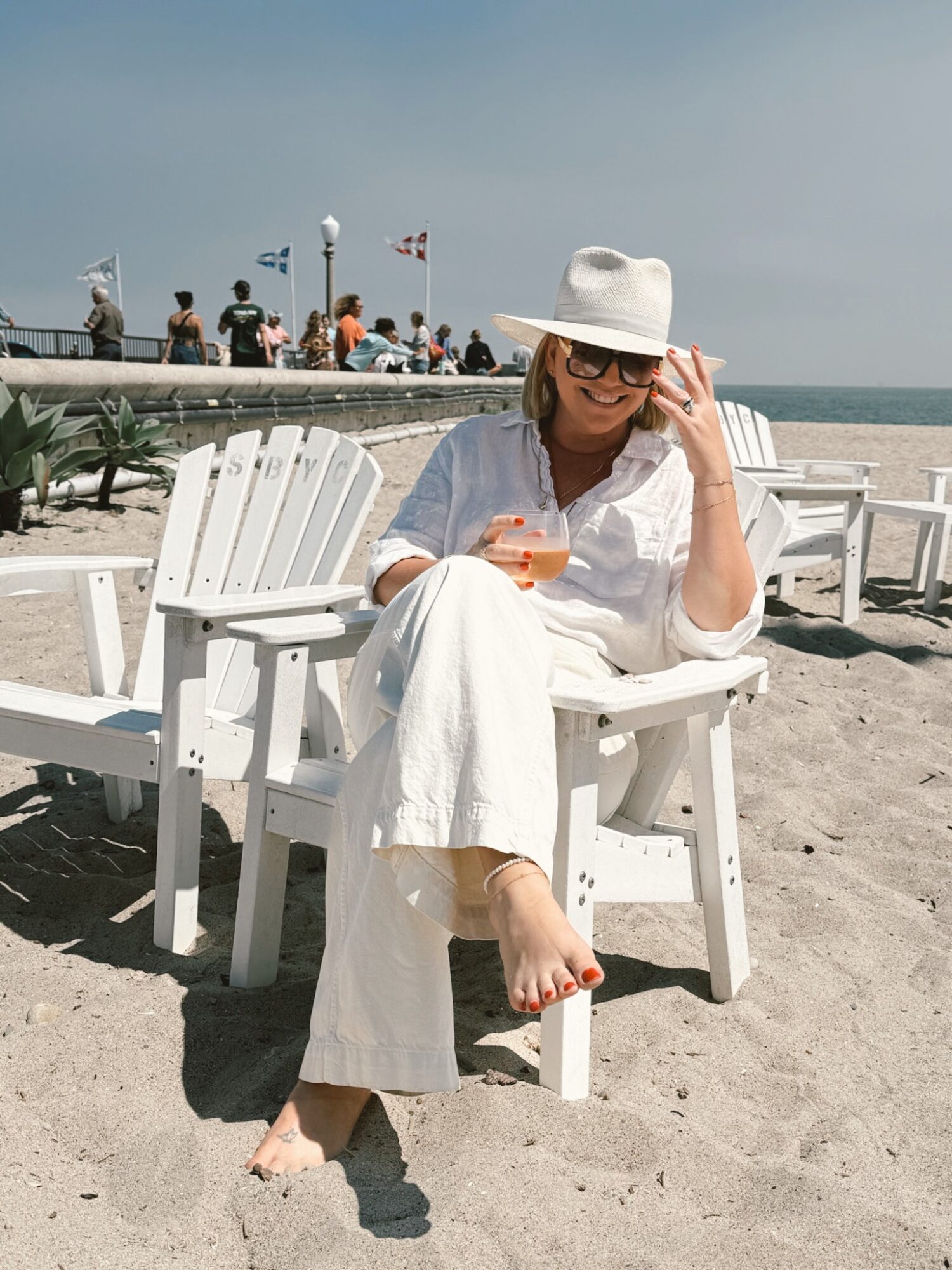 Woman sitting on a white beach chair, wearing a white hat and sunglasses, holding a drink, with a beach and pier in background.