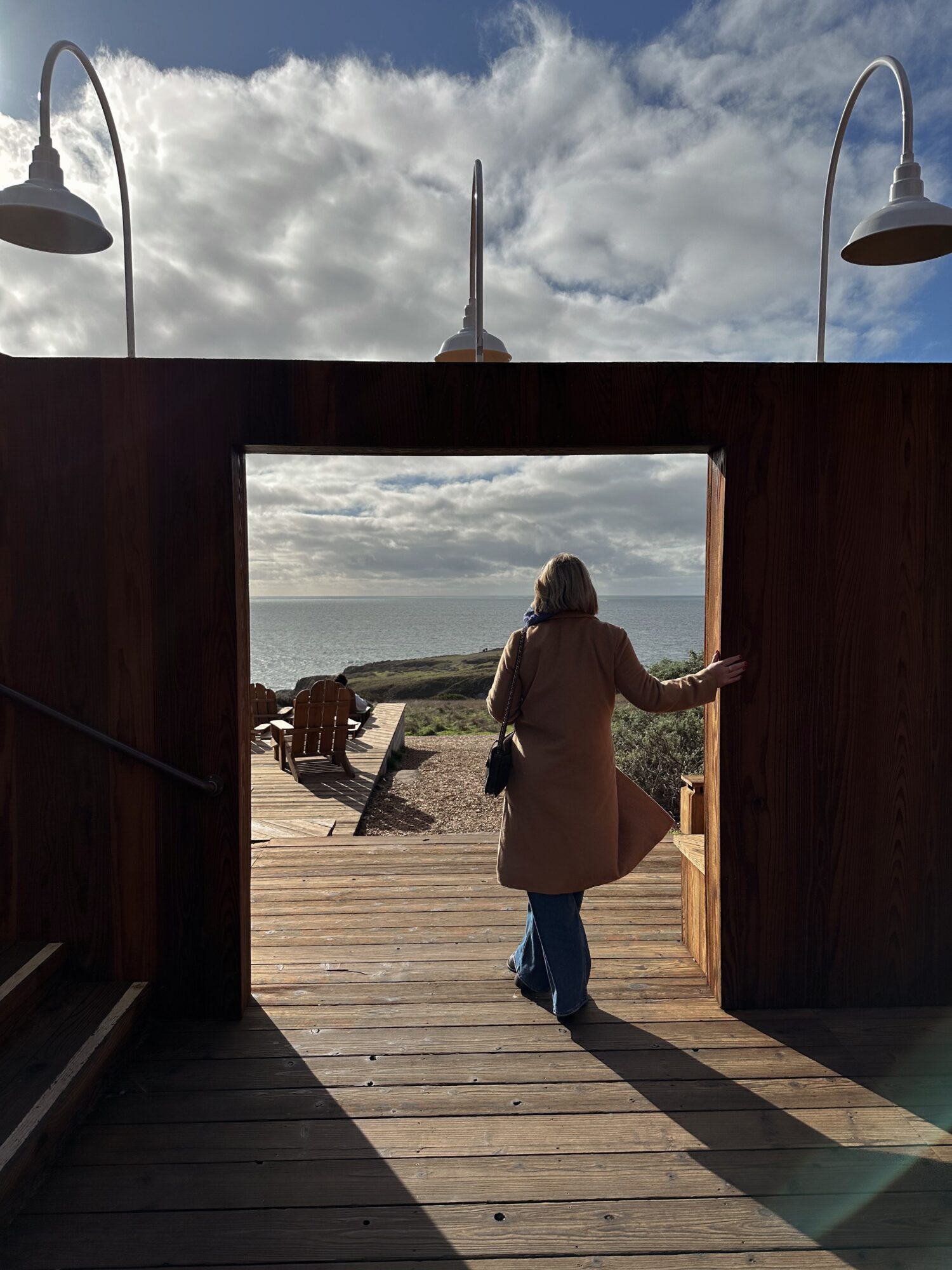 Person in coat walking out of a wooden doorway onto a deck with a view of sky and landscape, two lamps above.