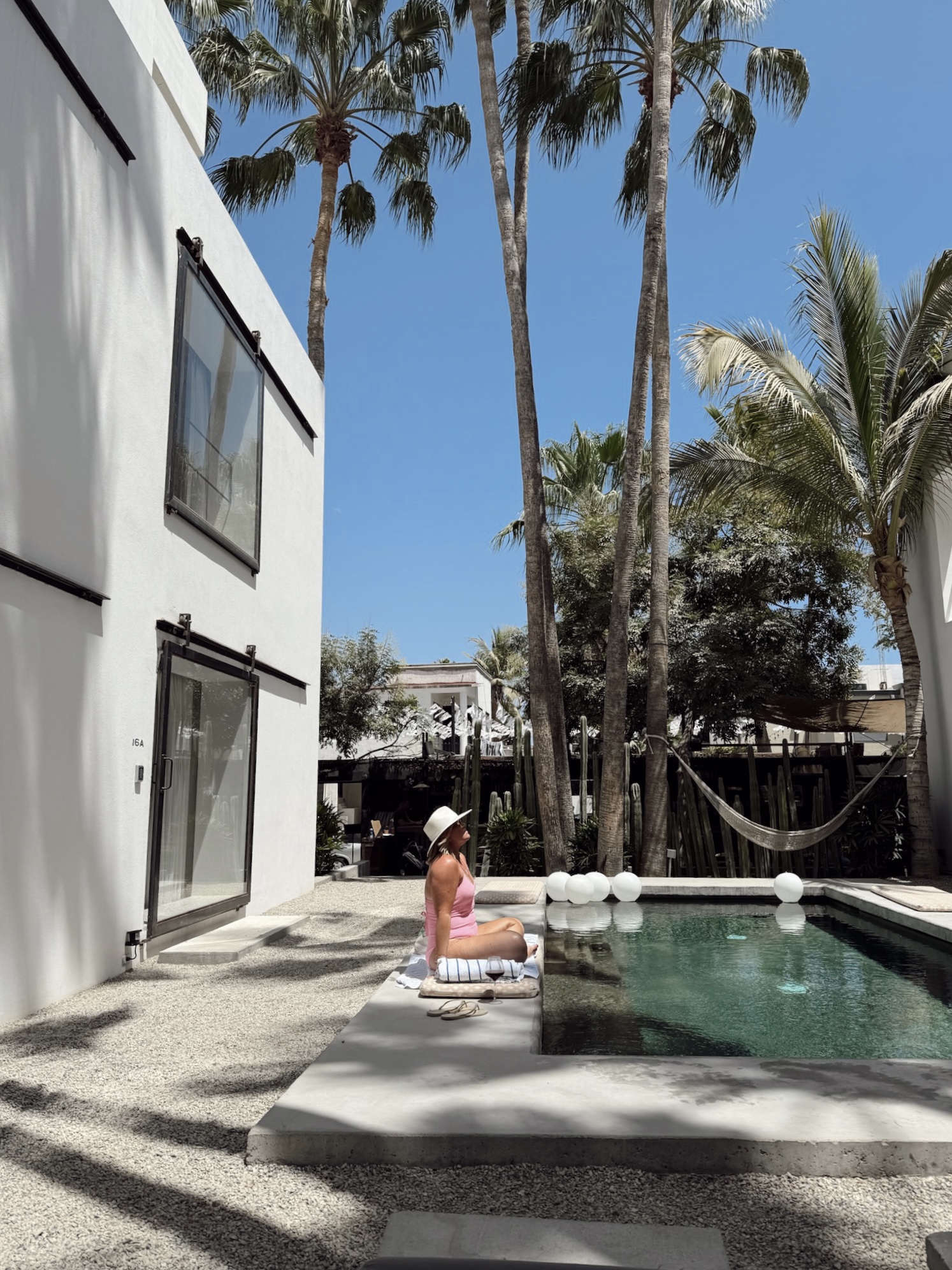 Person sitting by a swimming pool with tall palm trees and a white building in the background.