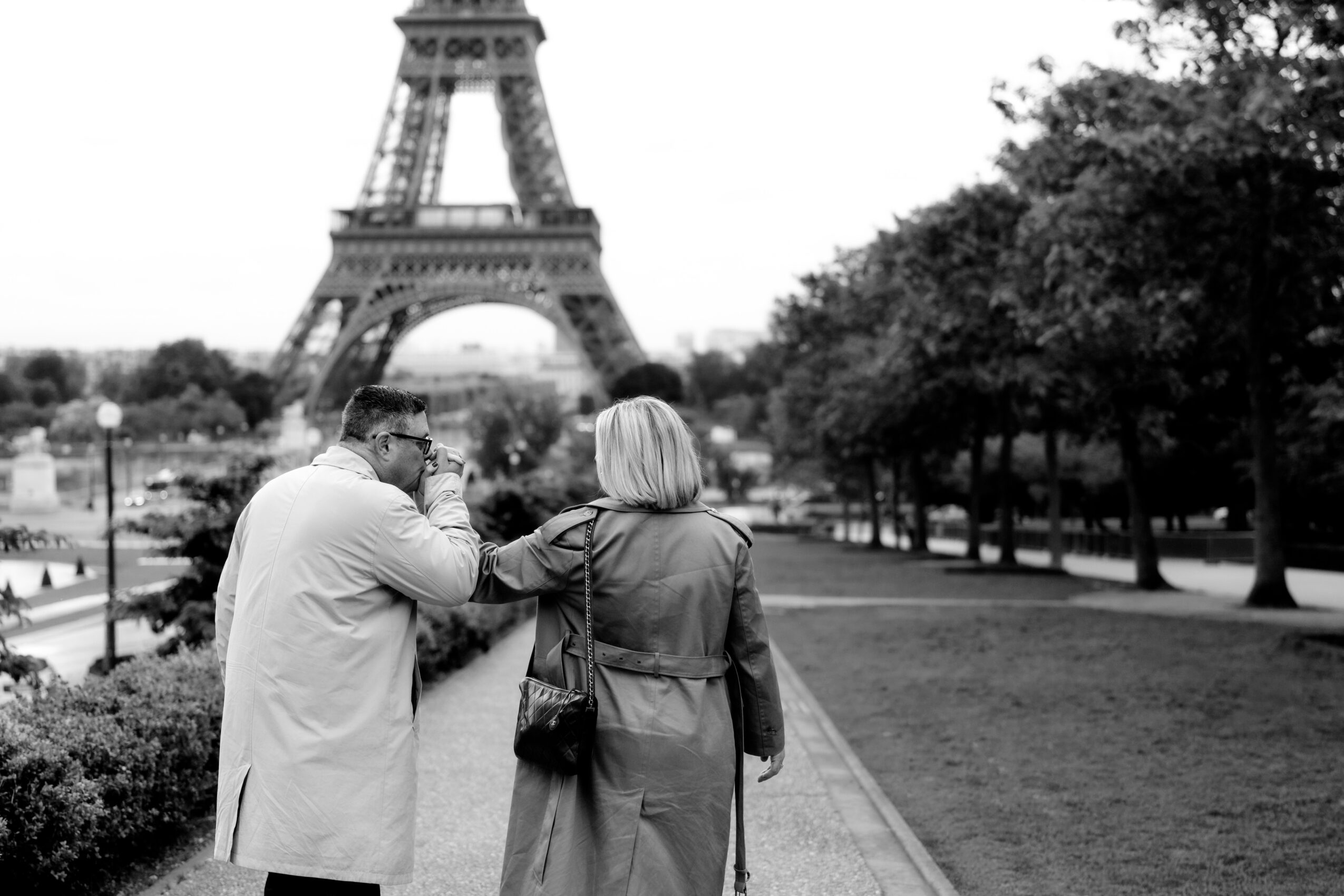 Two people walking near the Eiffel Tower, one with a coat and the other with a bag, in a park.