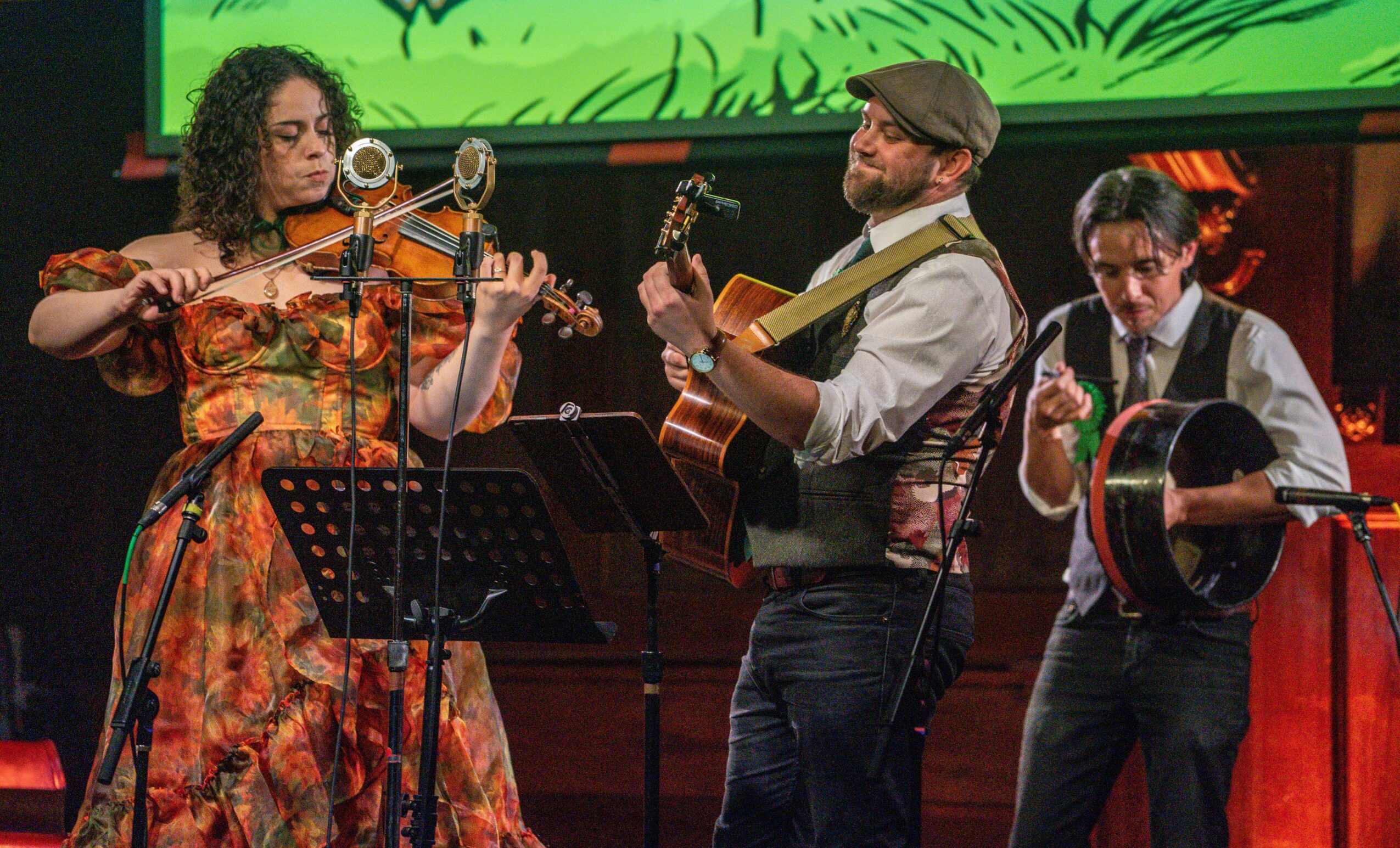 Three musicians perform on stage with instruments, including a violin, guitar, and tambourine, in front of a colorful background.