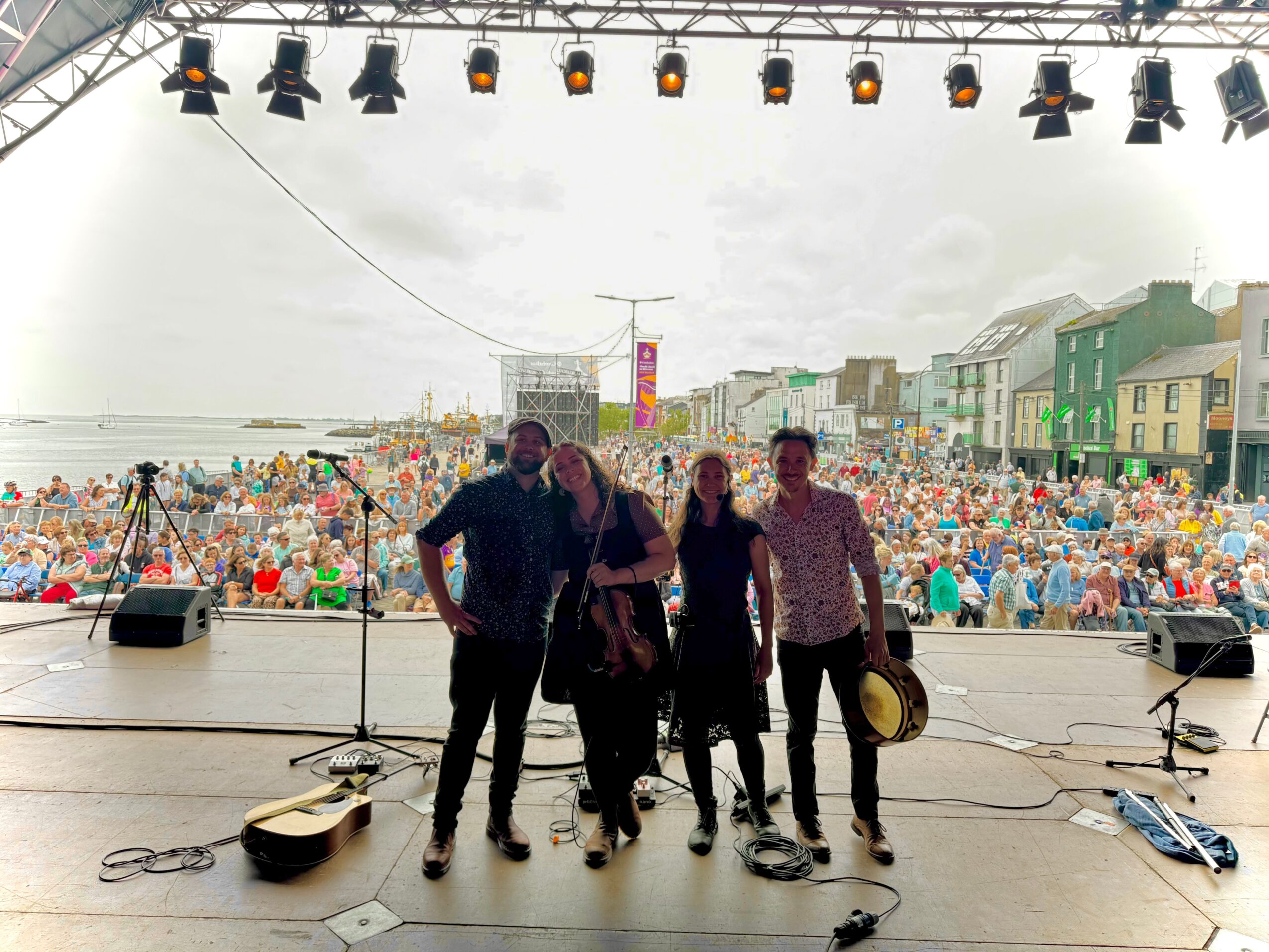 Four people stand on stage with a large crowd and buildings in the background, stage lights above, and musical instruments on stage.