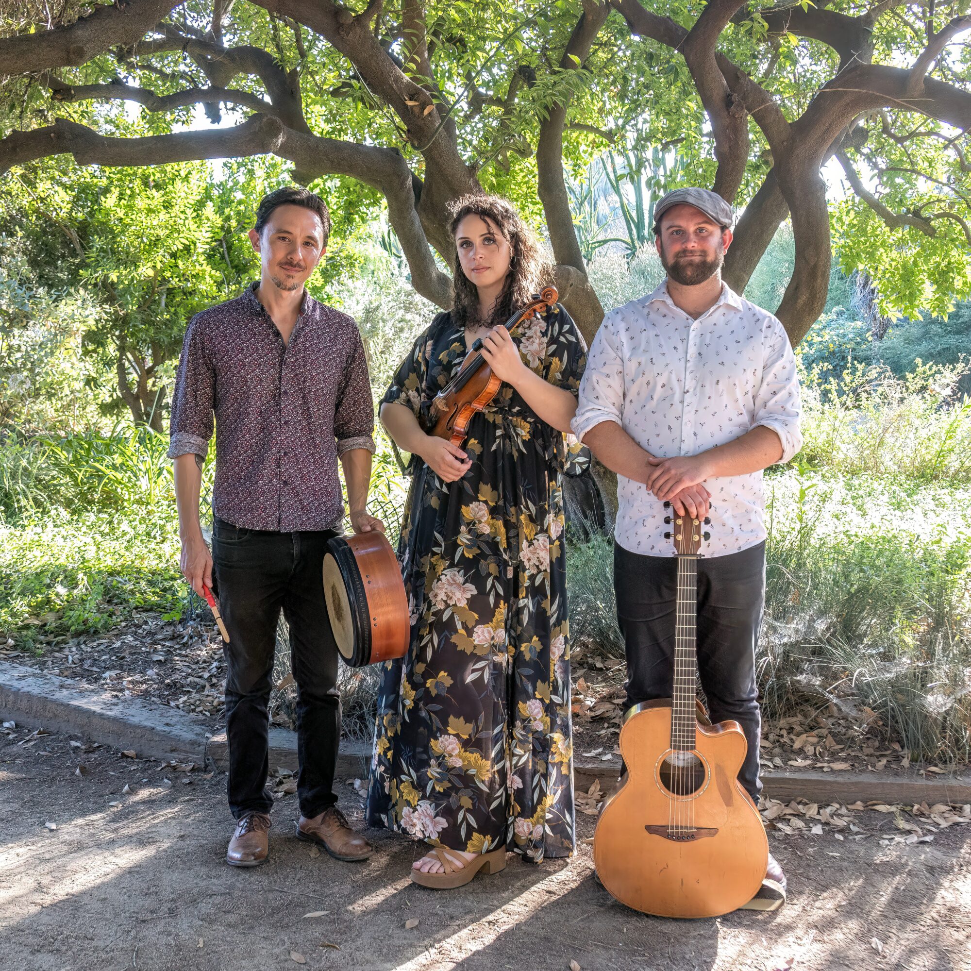 Three people standing outdoors under a large tree, two holding guitars, with greenery in the background.