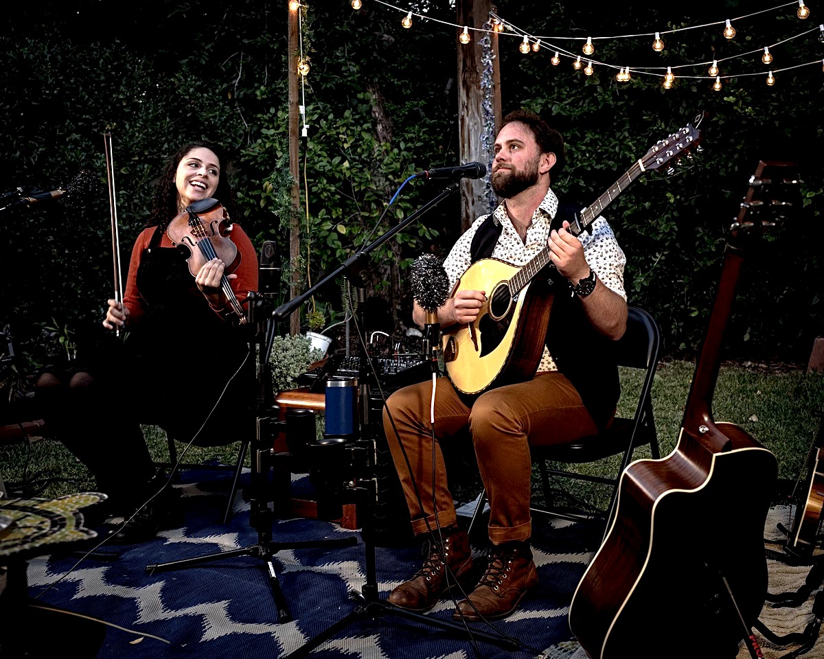 Two musicians perform outdoors at night, one singing and playing guitar, the other playing guitar, with string lights overhead.