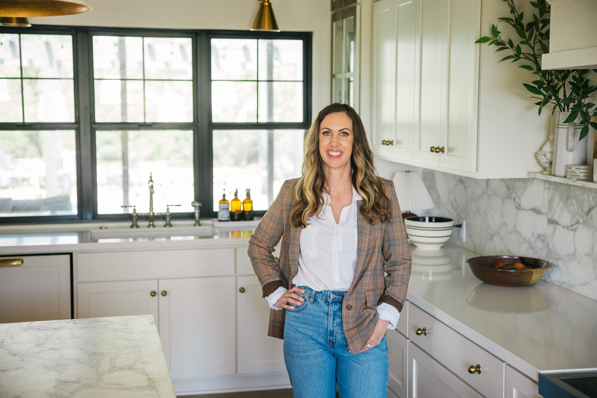 Woman standing in a bright kitchen with large windows, white cabinets, and marble countertops, smiling with hands in pockets.