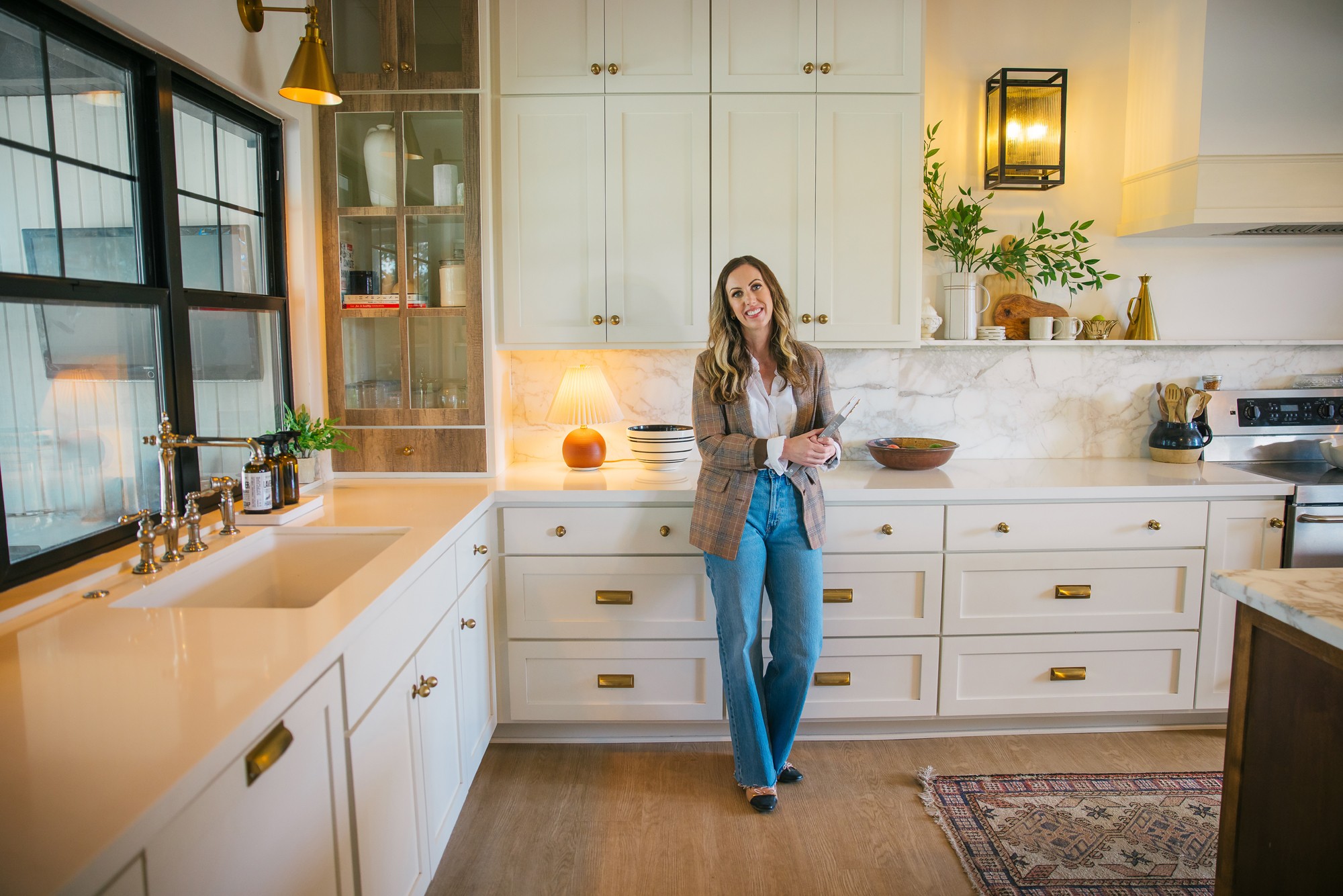Woman standing in a bright kitchen with white cabinets, wooden accents, and large windows, holding a cup.