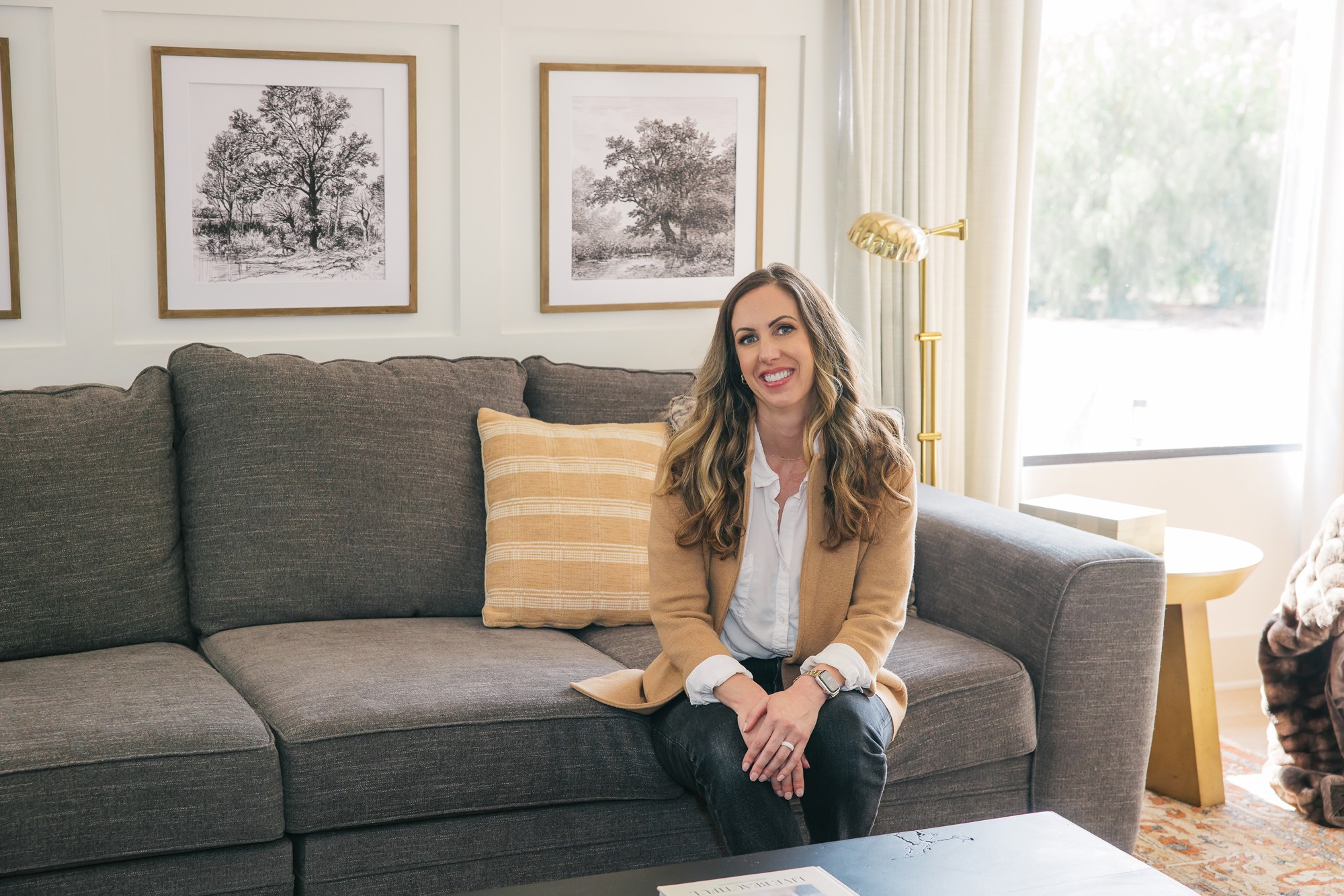 Woman sitting on a dark gray sofa in a living room, smiling, with framed pictures on the wall behind her.