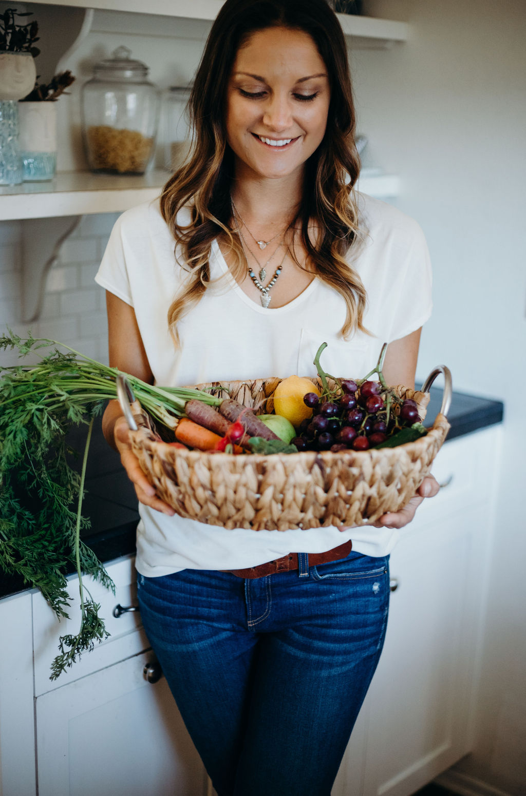 Woman holding a basket of fresh vegetables and fruits in a kitchen, smiling, wearing a white shirt and jeans.