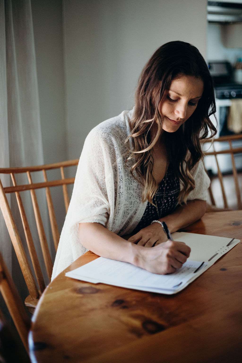 Woman with long hair writing on paper at a wooden table in a room with natural light.