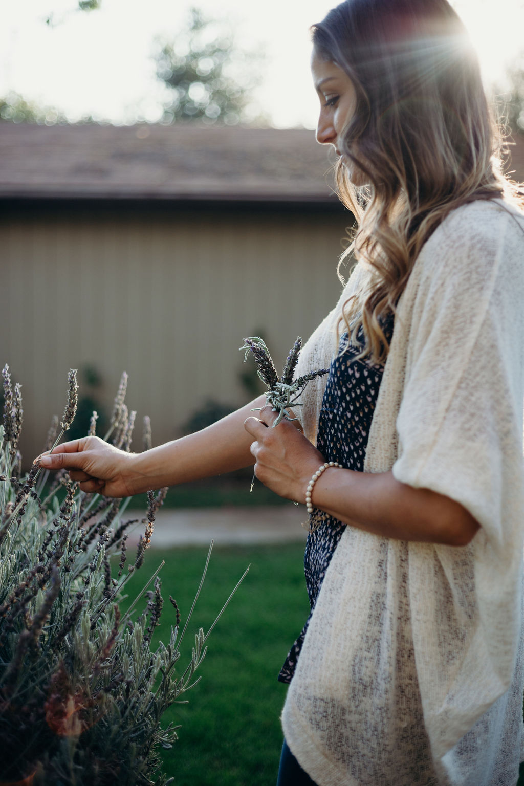 Young woman with long hair gently touching lavender plants outdoors, sunlight shining behind her, in a garden setting.